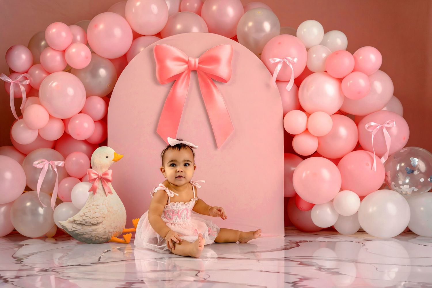 Pink and white balloon arch with decorative bow creates elegant backdrop for first birthday celebration photoshoot.