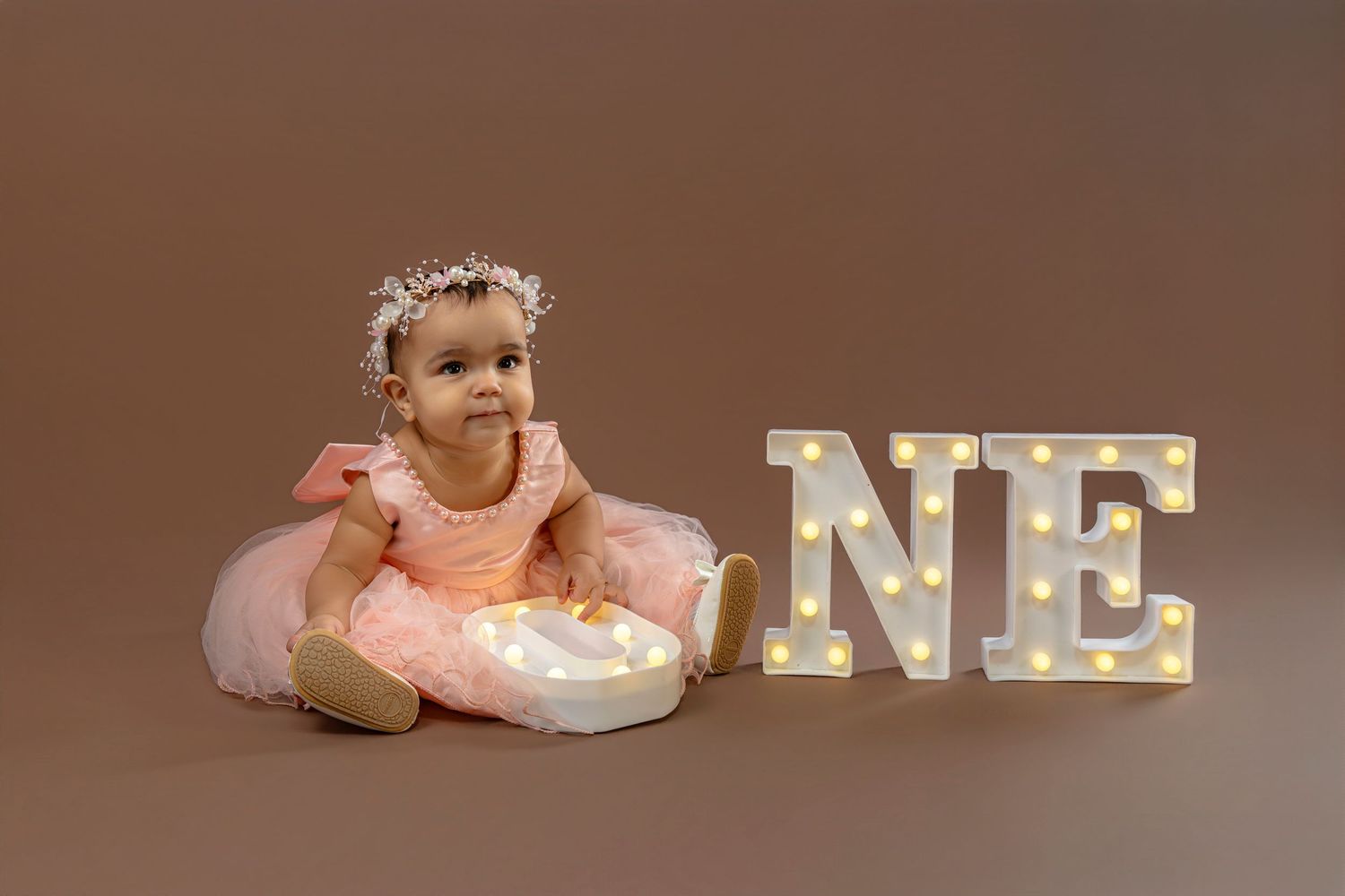Baby in pink tutu dress sits beside illuminated marquee letter 'ONE' on brown backdrop during first birthday photoshoot.