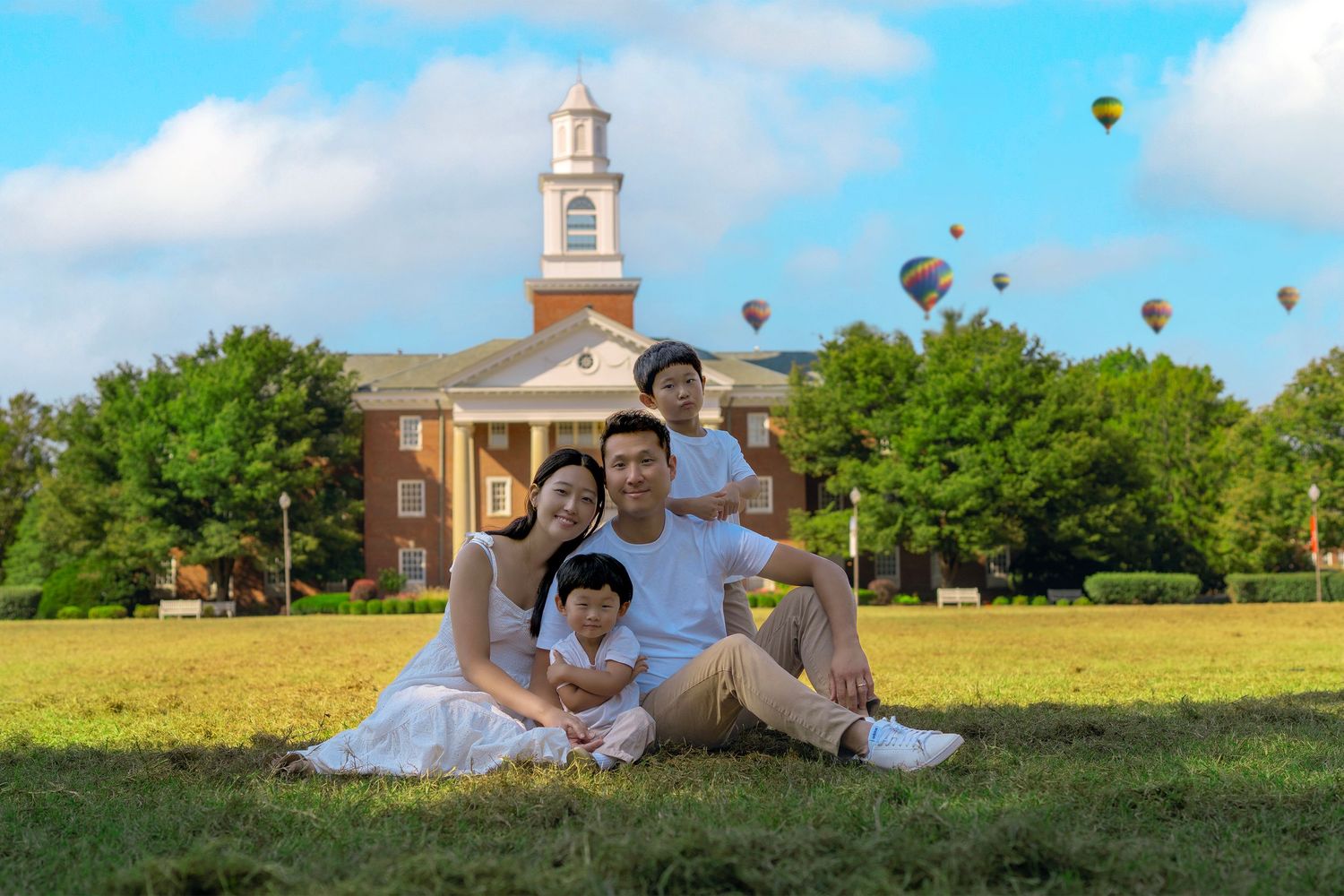 Asian family of four sitting on grass before a brick building with colorful hot air balloons in blue sky.