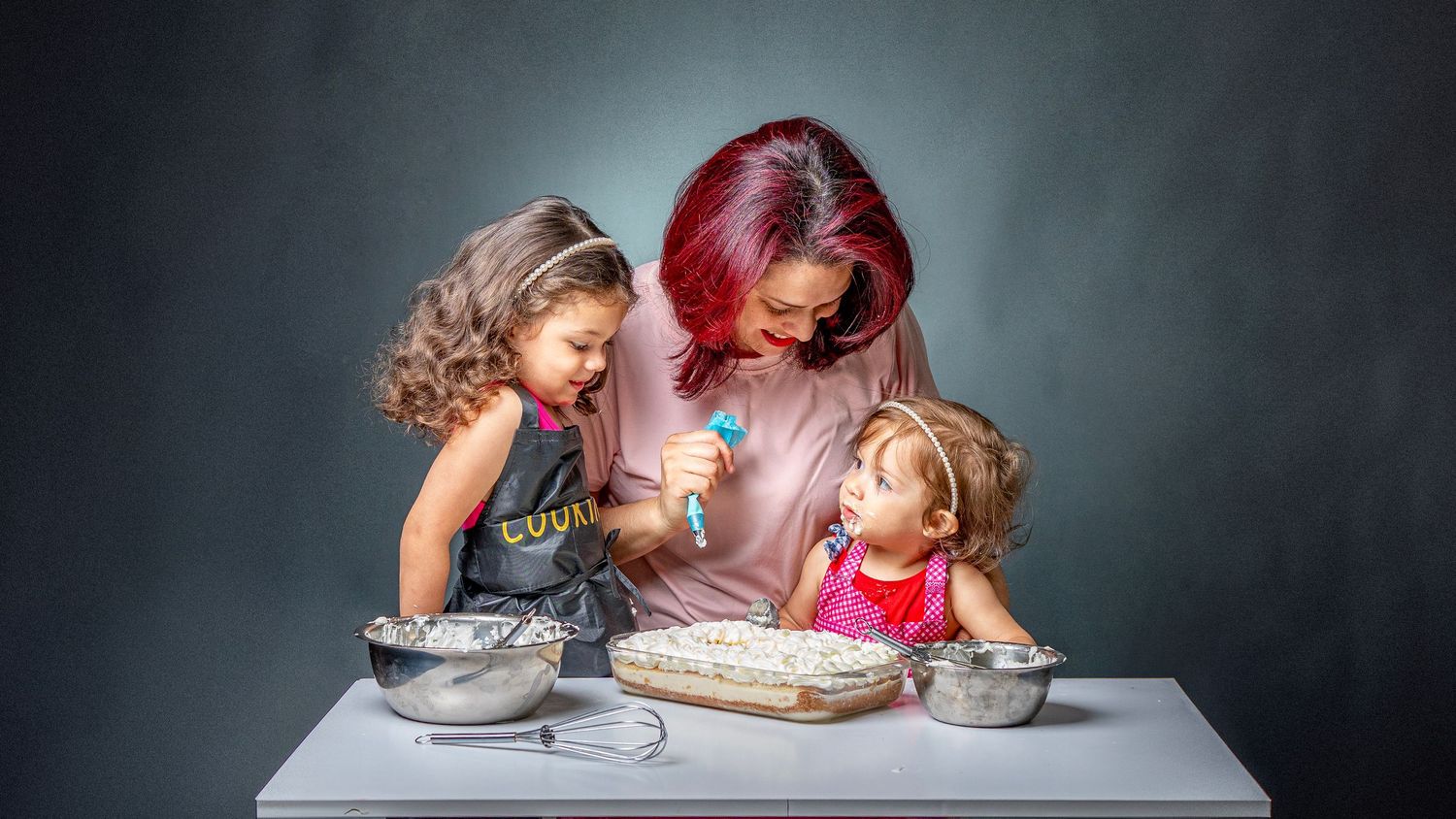 A red-haired woman and two young girls baking together at a white table with mixing bowls and a baking dish.