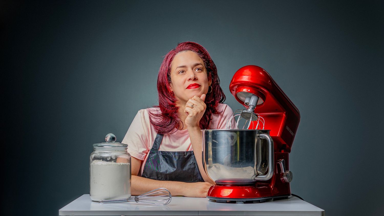 Woman with pink hair sits pensively at a table with a red stand mixer and flour jar in a kitchen setting.