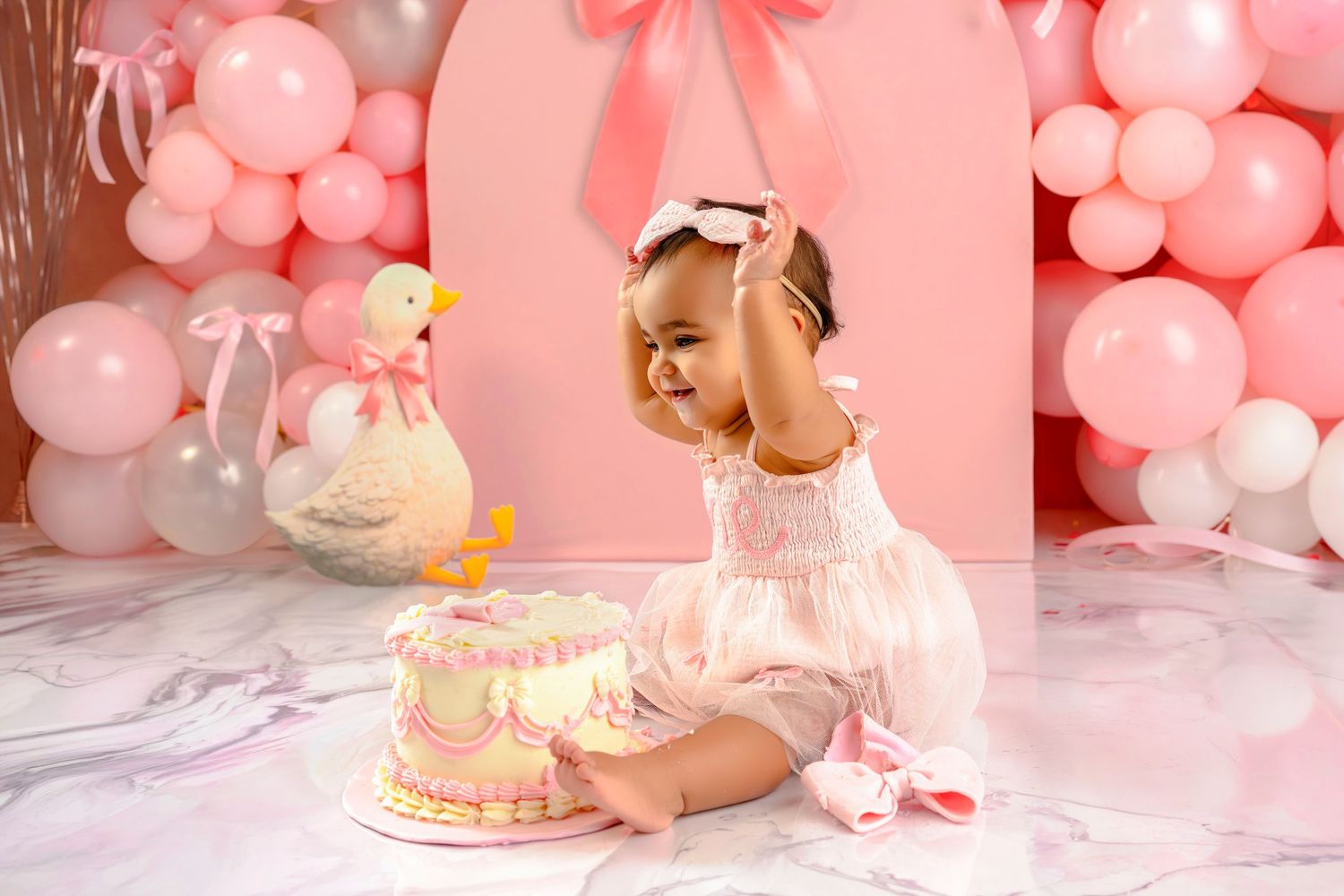 Pink balloon decorations surround a white and pink birthday cake with a number one candle on top.