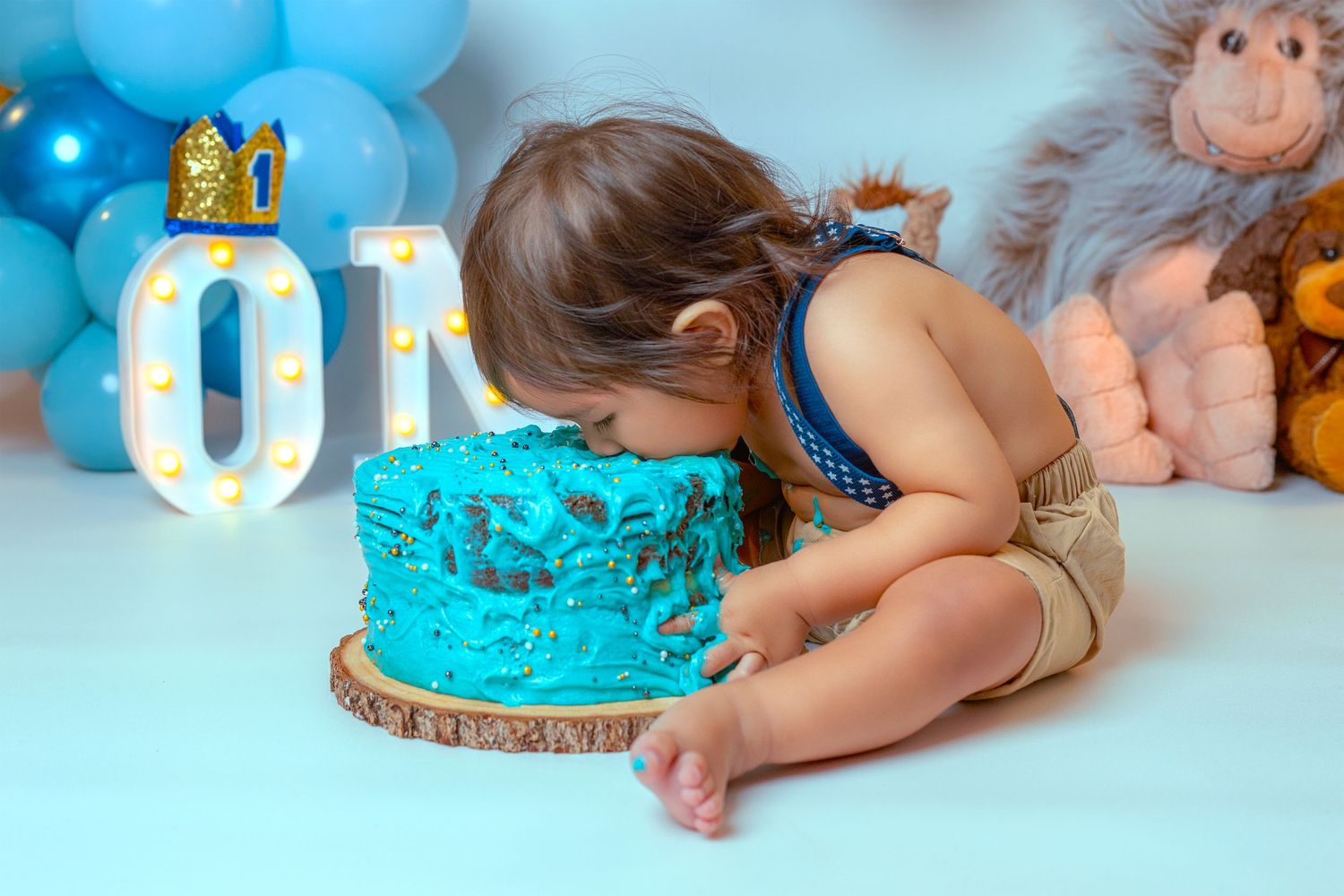Child in blue tank top leans over to bite turquoise birthday cake with number one candle and blue balloon decorations.