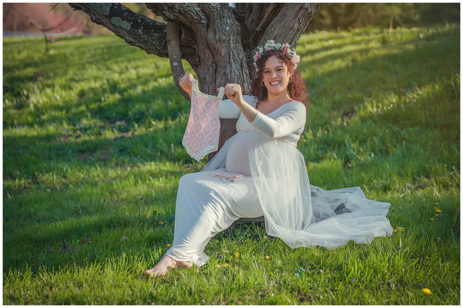 Pregnant woman in white dress sitting under tree holding pink baby clothes in sunny green park.