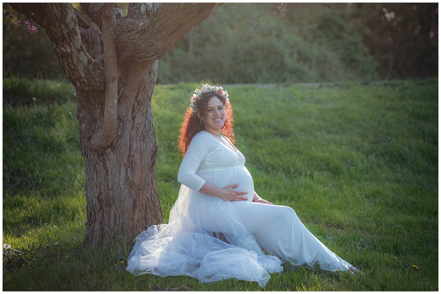 Pregnant woman in white tulle gown sitting by tree in golden hour light, cradling baby bump in lush green field.