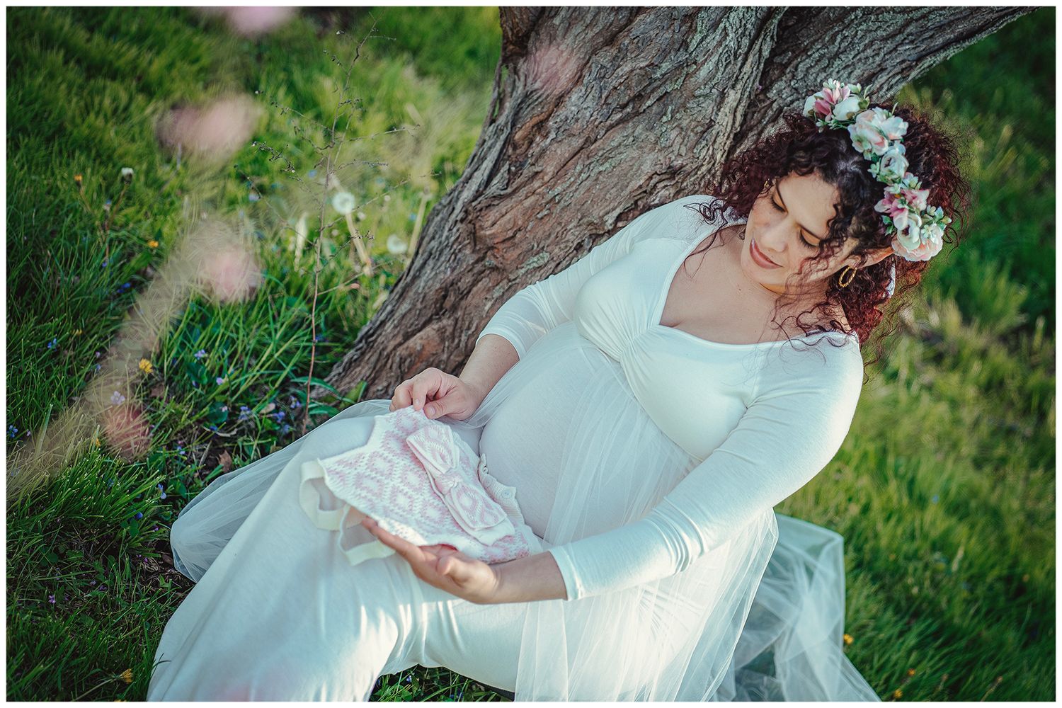 Pregnant woman in white floral crown dress sits against tree trunk holding delicate lace baby item.