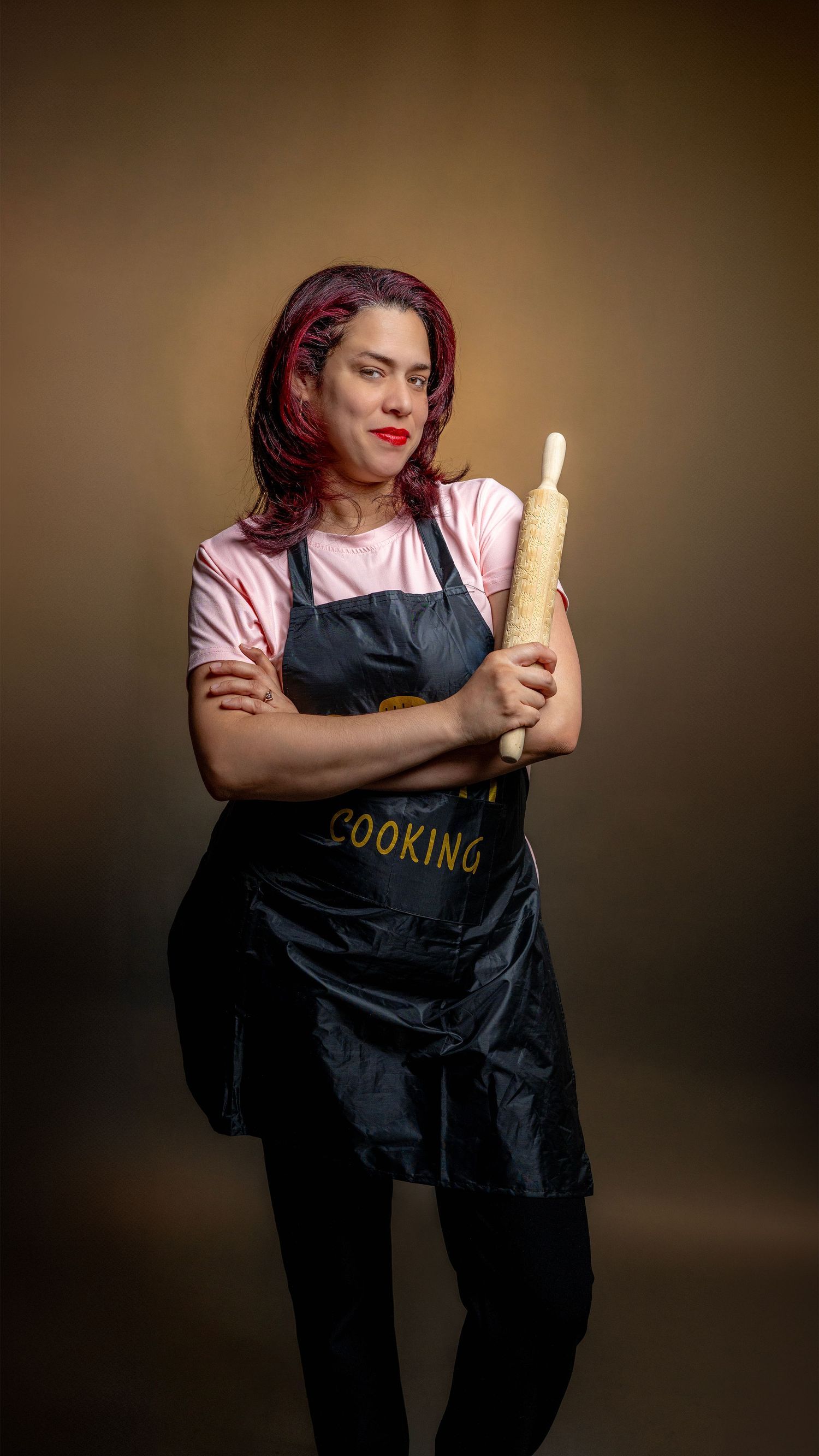Woman with red hair wearing a black cooking apron holds a rolling pin with a confident expression against a brown background.