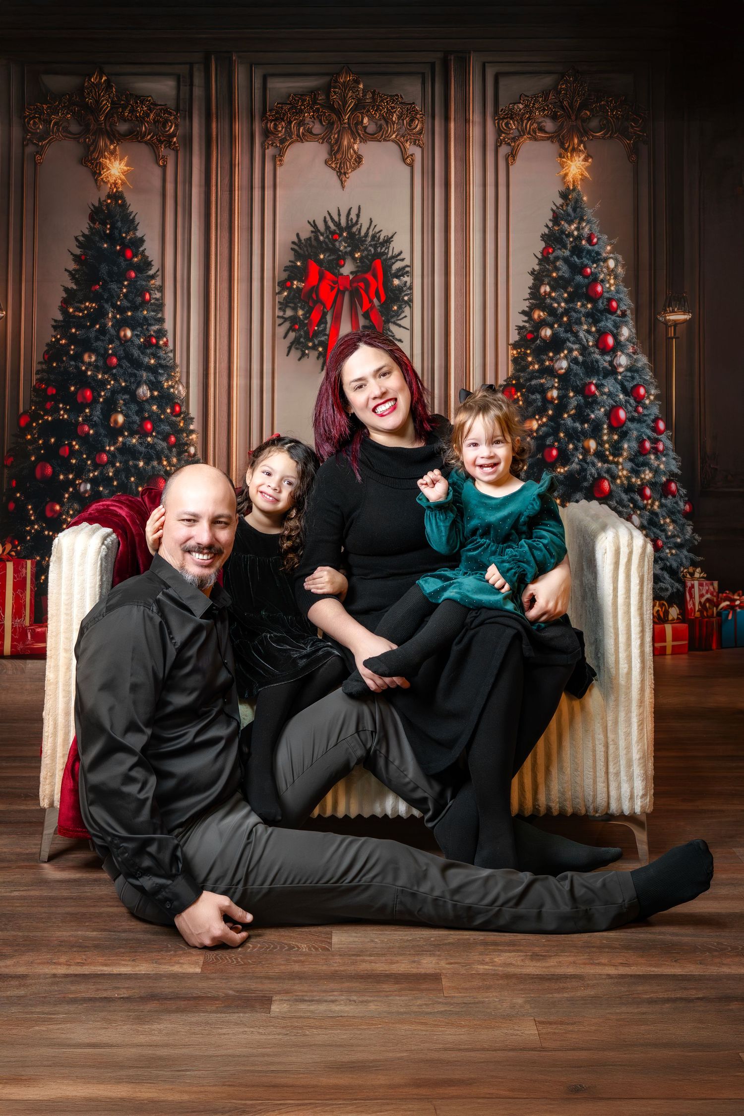 Family of four posing joyfully in front of decorated Christmas trees in an elegant dark wood-paneled room.