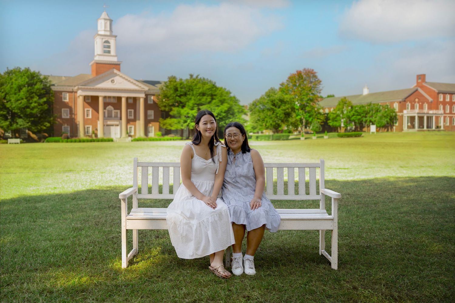 Two women sitting together on a white bench on a green lawn in front of a brick university campus building.