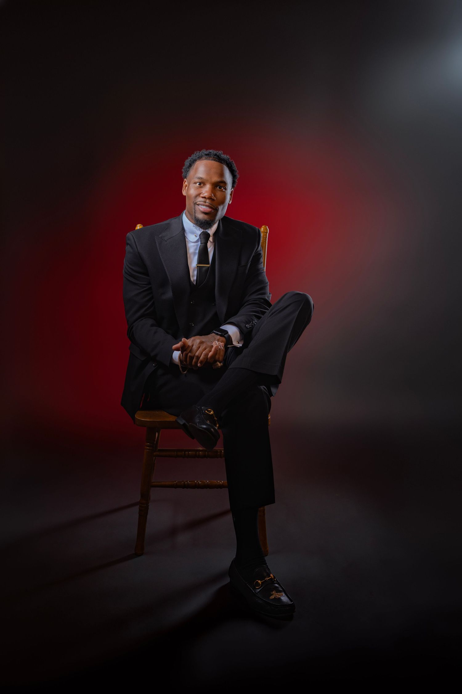 A man in a dark suit sits confidently on a wooden chair against a dramatic red and black studio background.