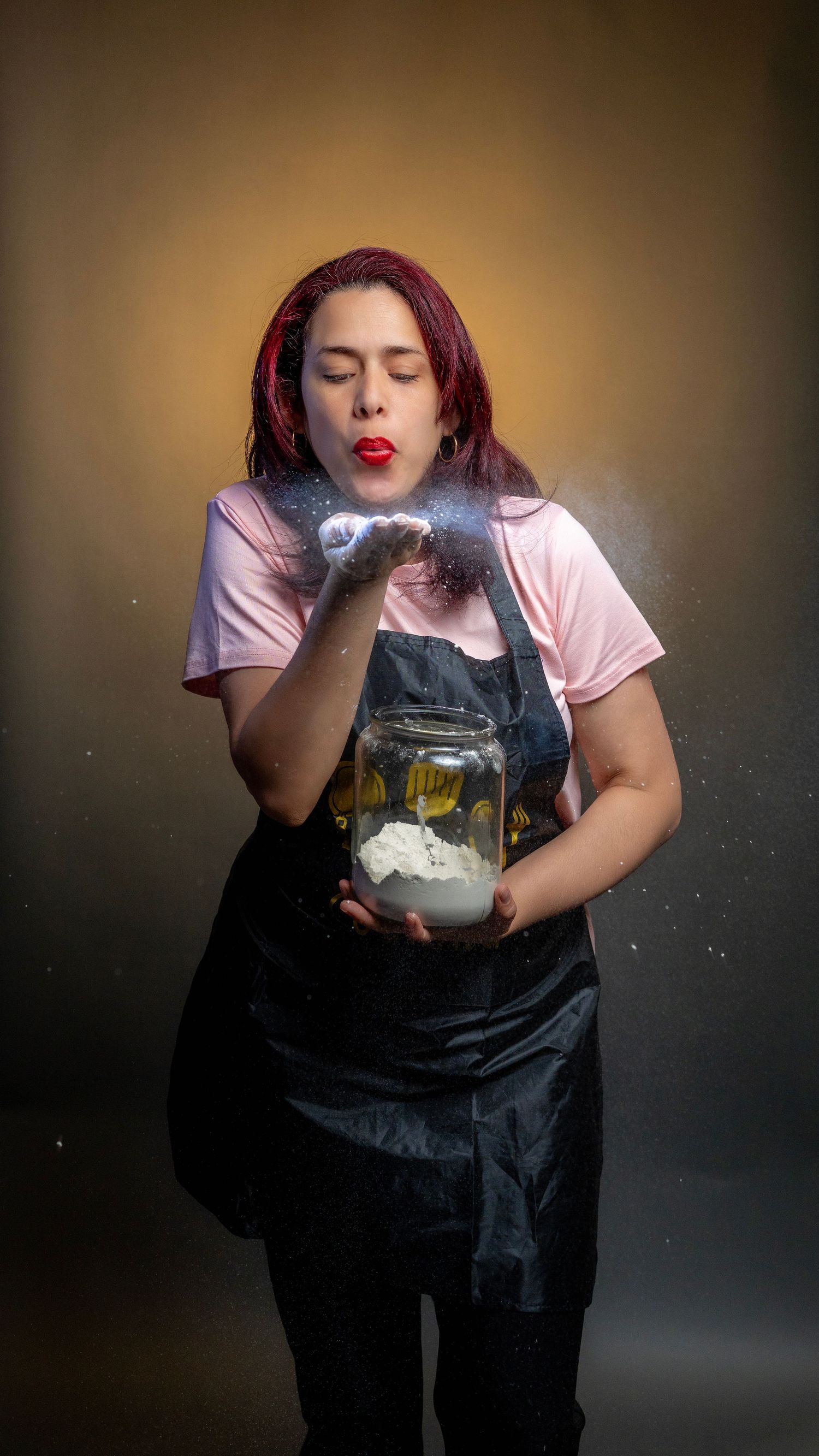 Woman with red hair blowing glittery powder from hands while holding a jar, wearing a black apron over pink shirt.