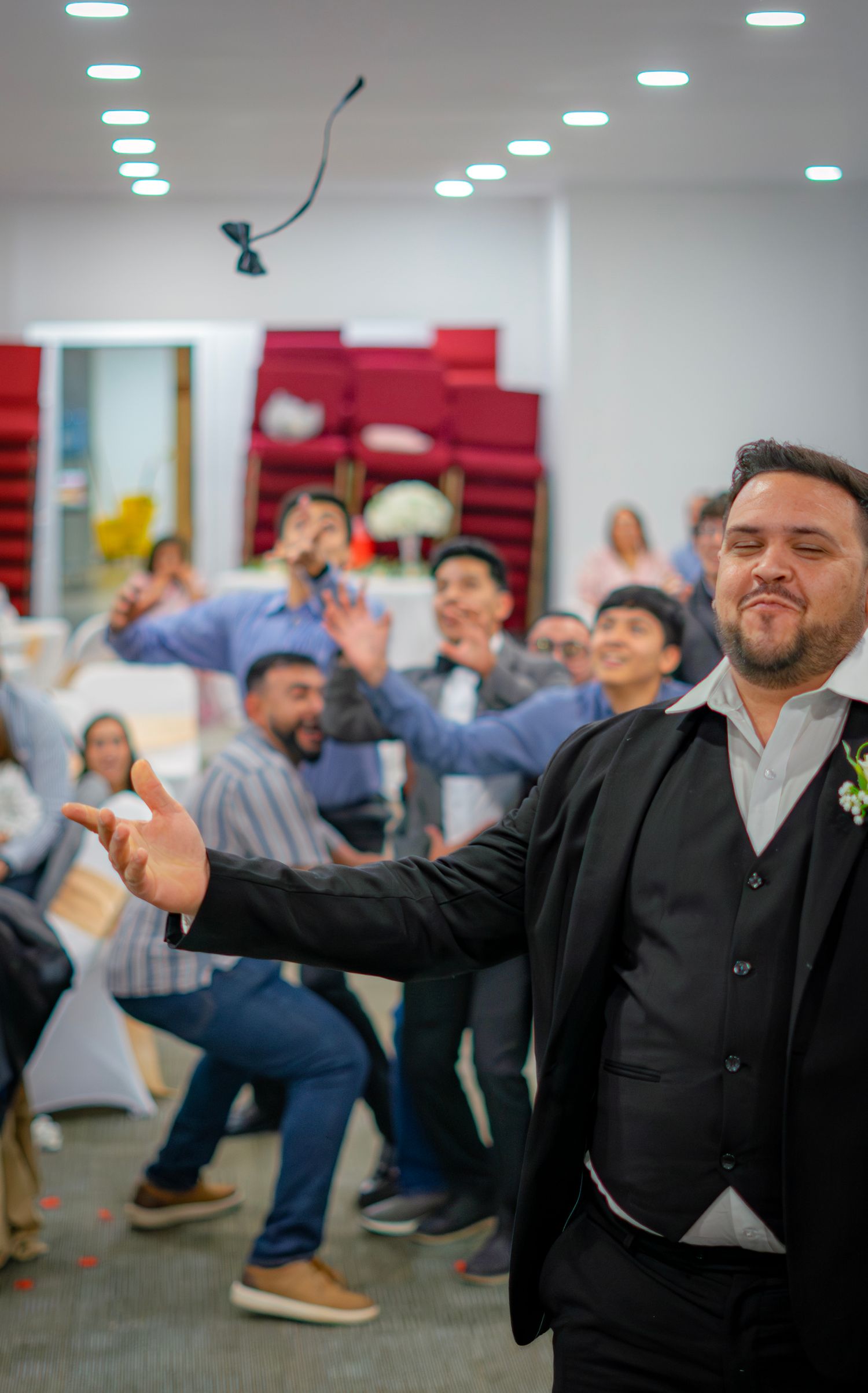 Groom in black suit tossing bouquet to excited wedding guests in a decorated reception hall.