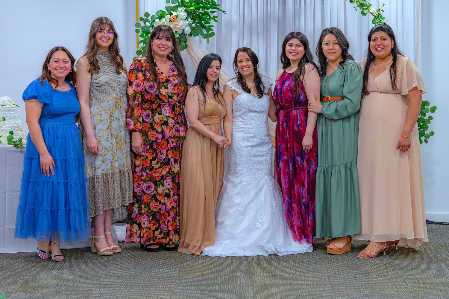 Group of eight women in colorful dresses posing together at a wedding celebration, with a bride in white gown at center.