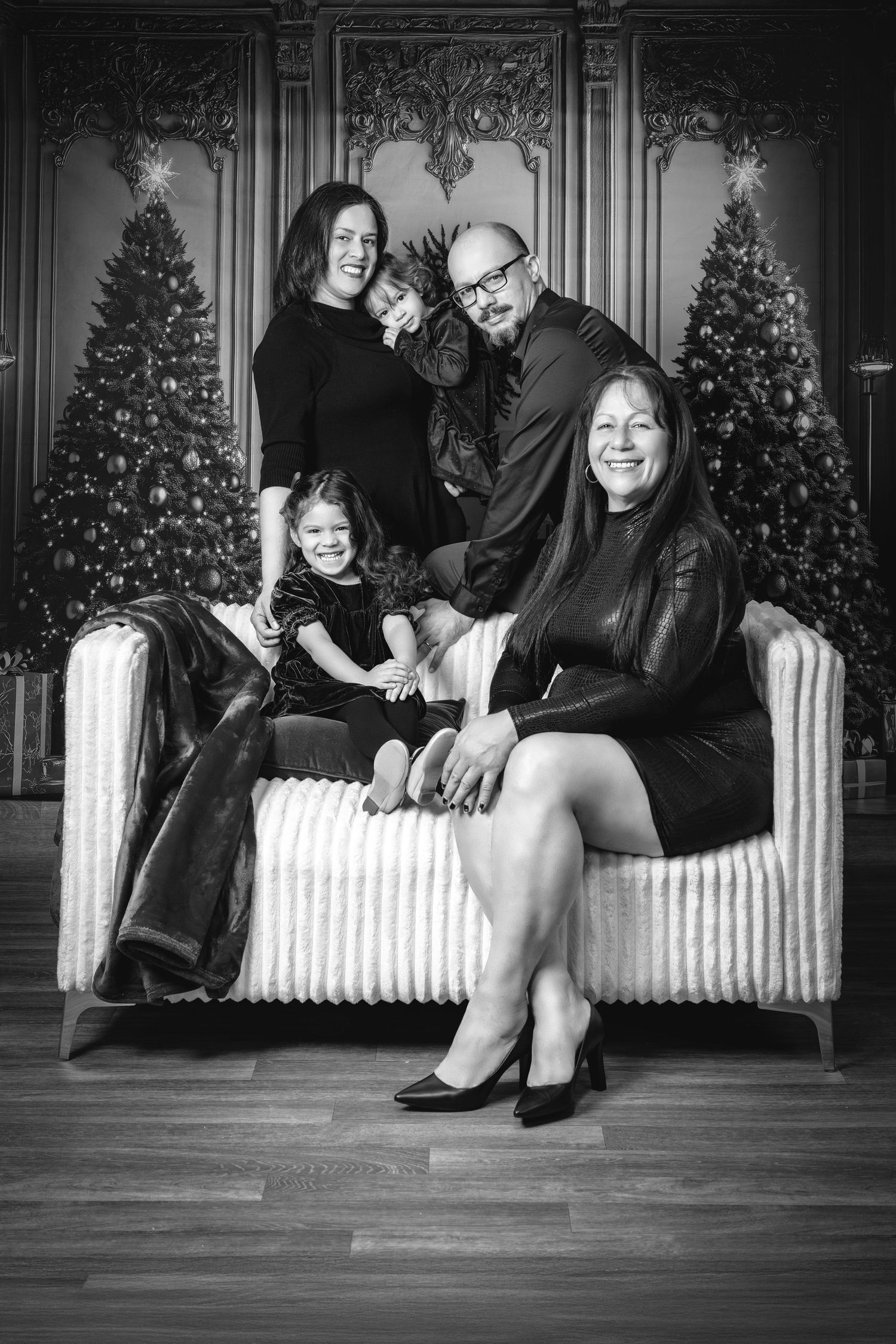 Black and white family portrait with two Christmas trees in background, sitting on striped couch in formal attire.