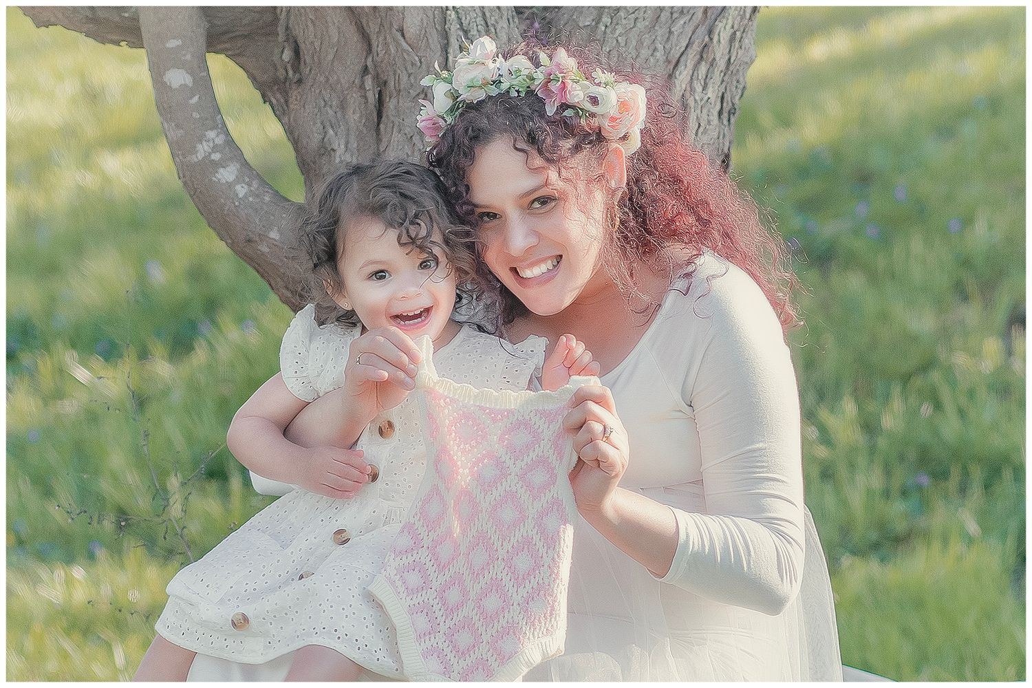 Mother and toddler daughter in white dresses with floral crowns smiling outdoors by a tree in a green meadow.
