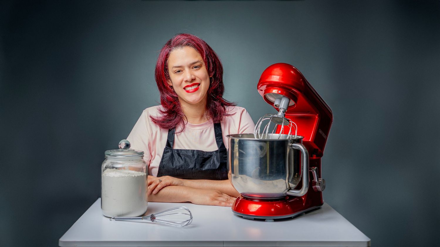 Woman with pink hair in apron smiling beside a red stand mixer and flour canister on white table.