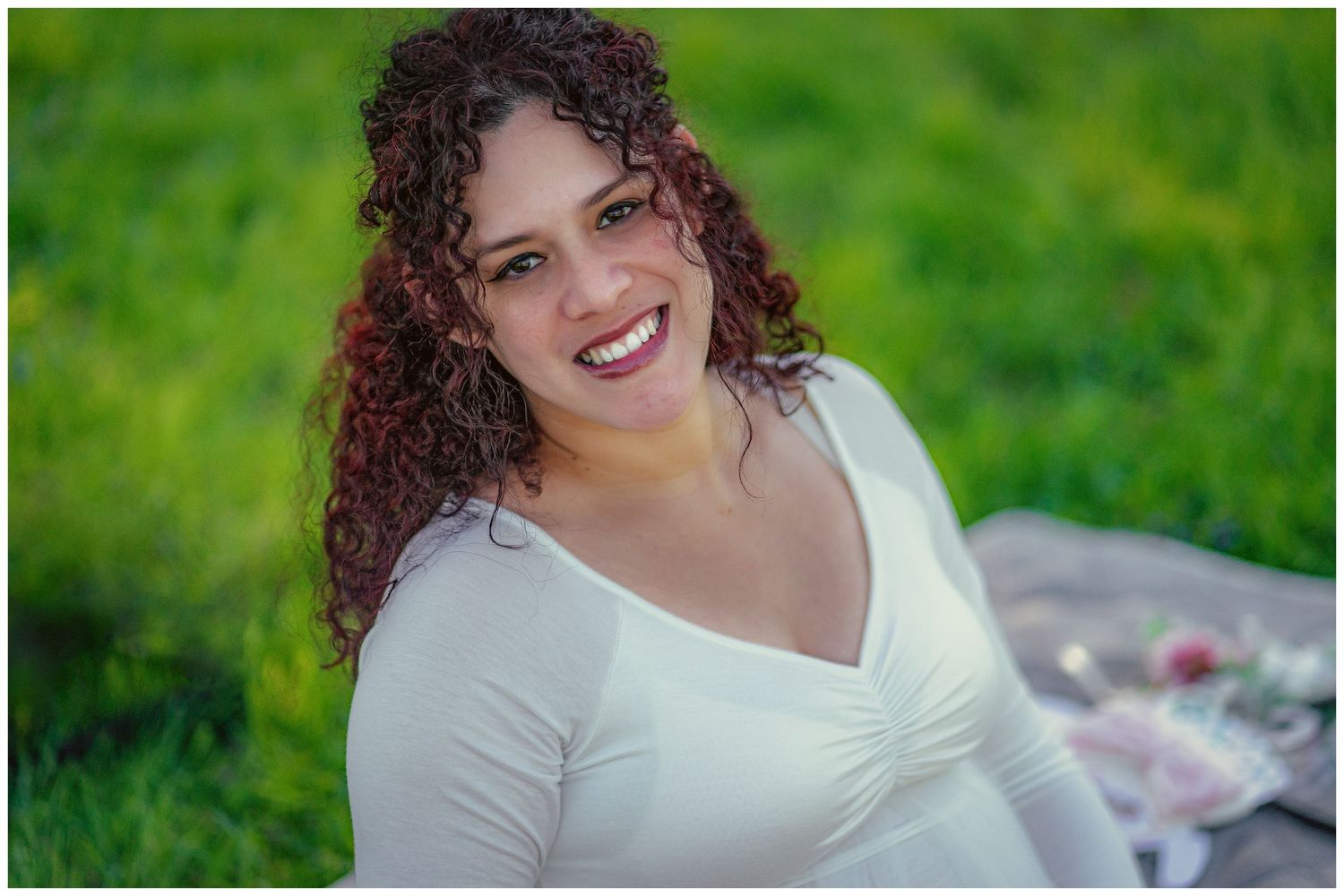 A smiling woman with curly hair wearing a white top sits outdoors on a blanket surrounded by lush green grass.