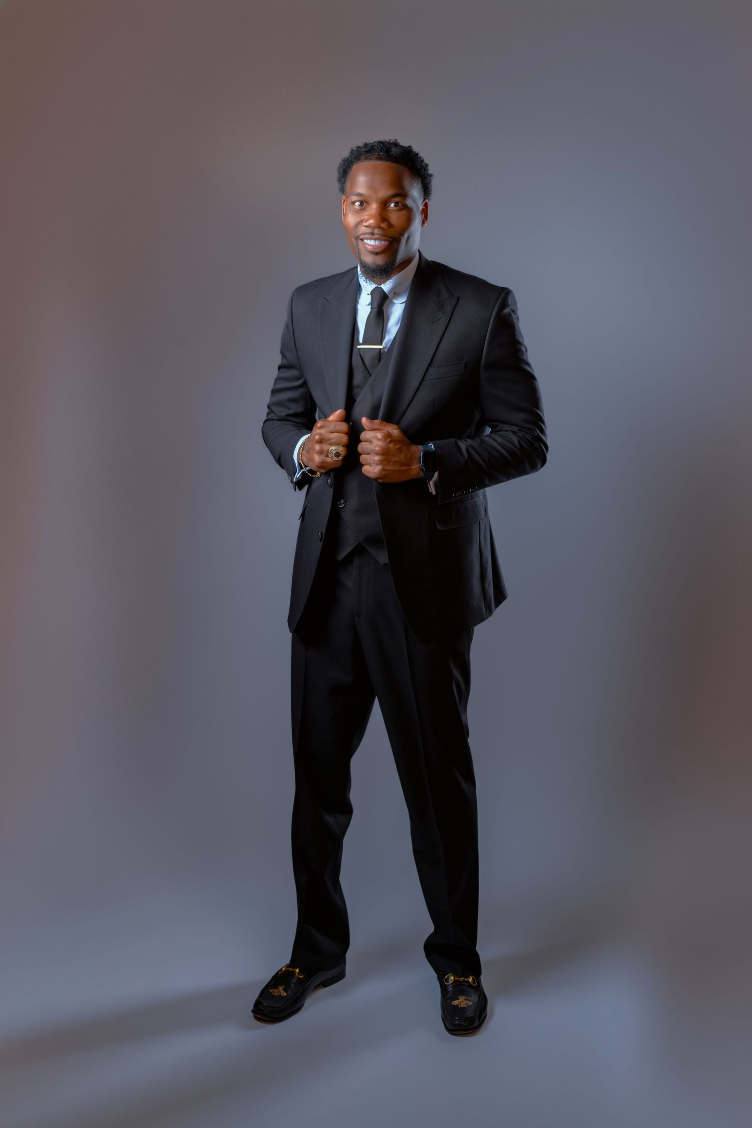 A confident Black man in a sharp black suit and tie poses against a gray background in a professional studio portrait.