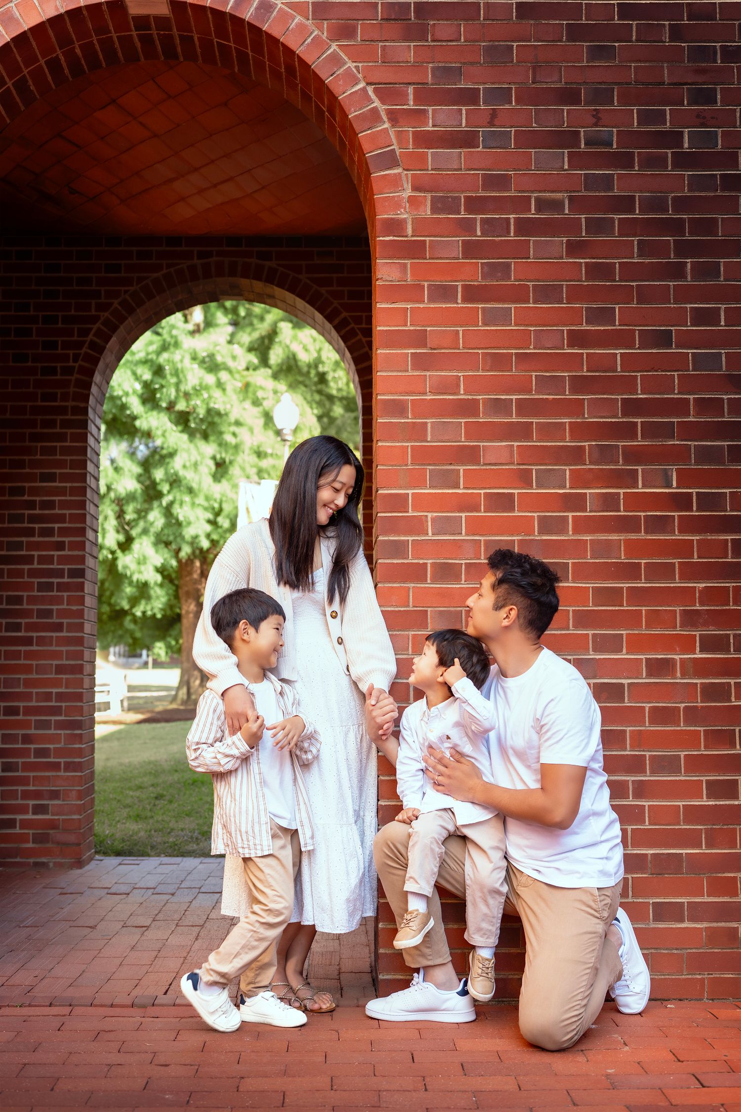 Happy Asian family with two young boys posing under a red brick arch in a park, mother in white dress appears pregnant.