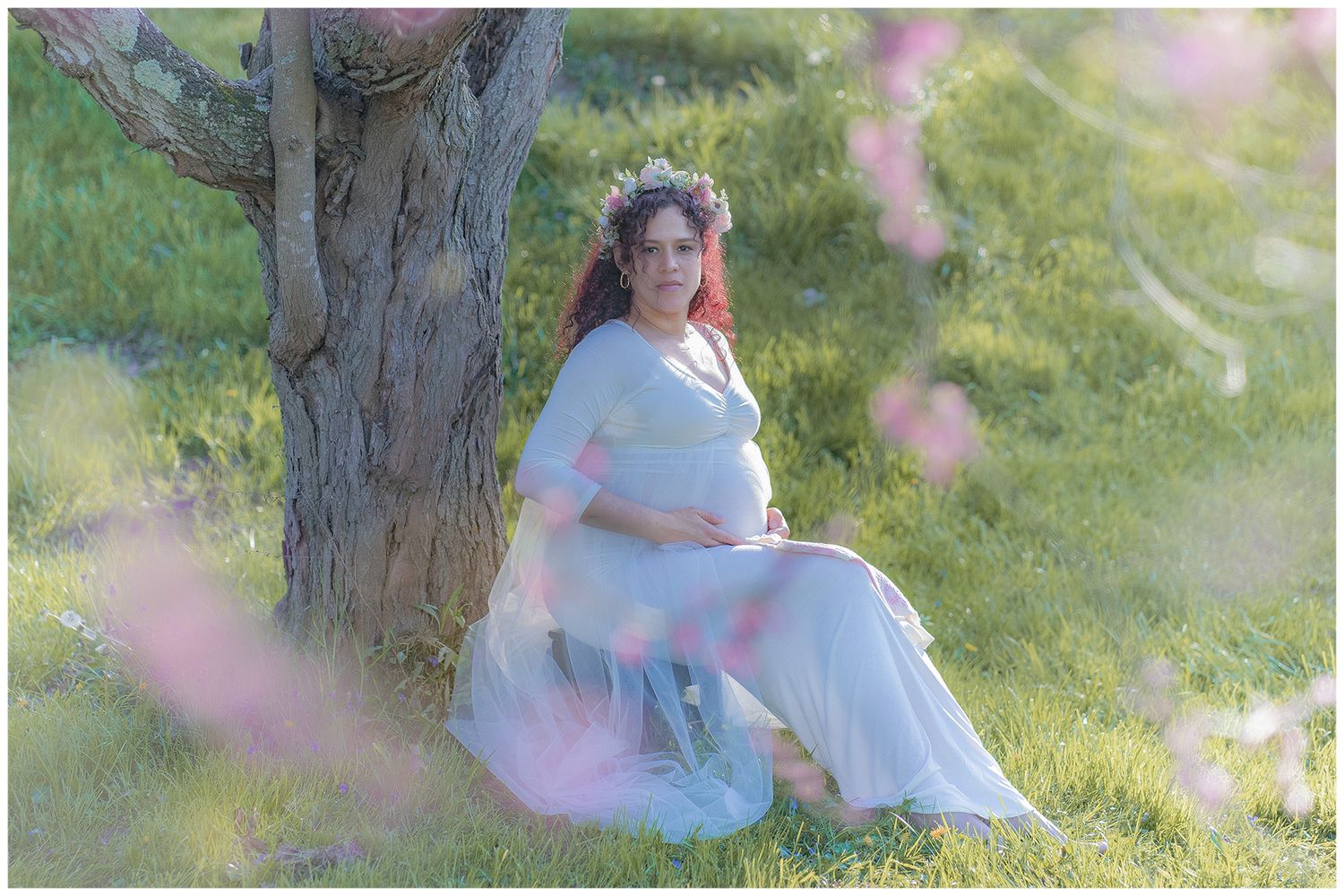 Pregnant woman in flowing blue gown leans against tree in blooming garden for a dreamy maternity photo session.