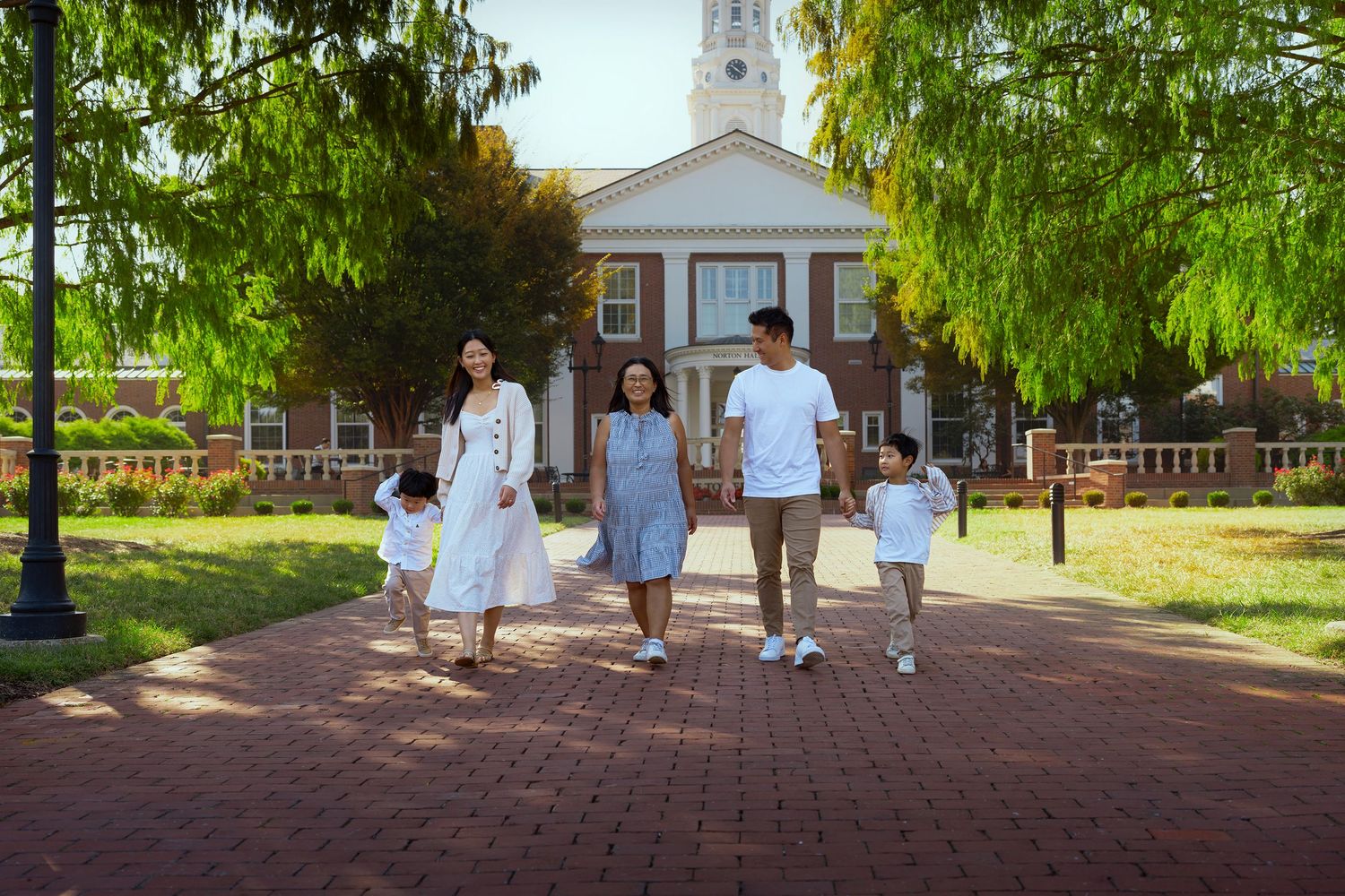 People walking on a brick path through a garden with willow trees and buildings in the background.