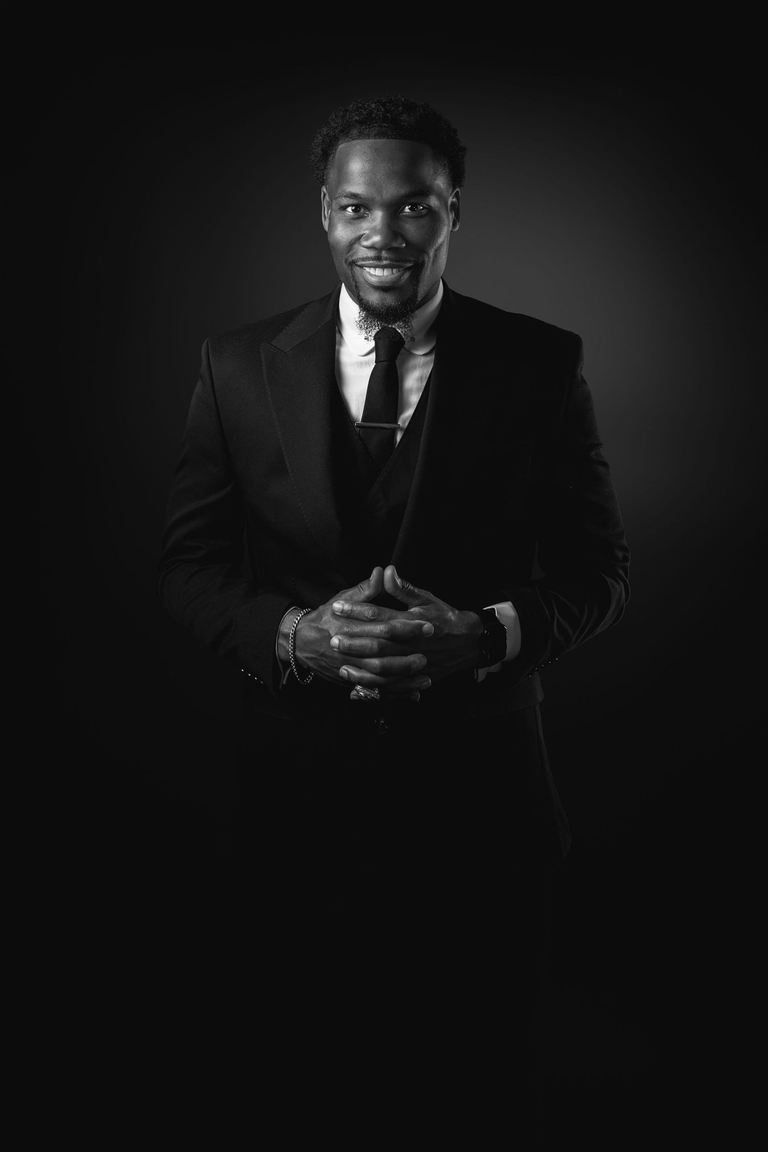 Professional black and white portrait of a person in a dark suit and tie smiling against a black background.