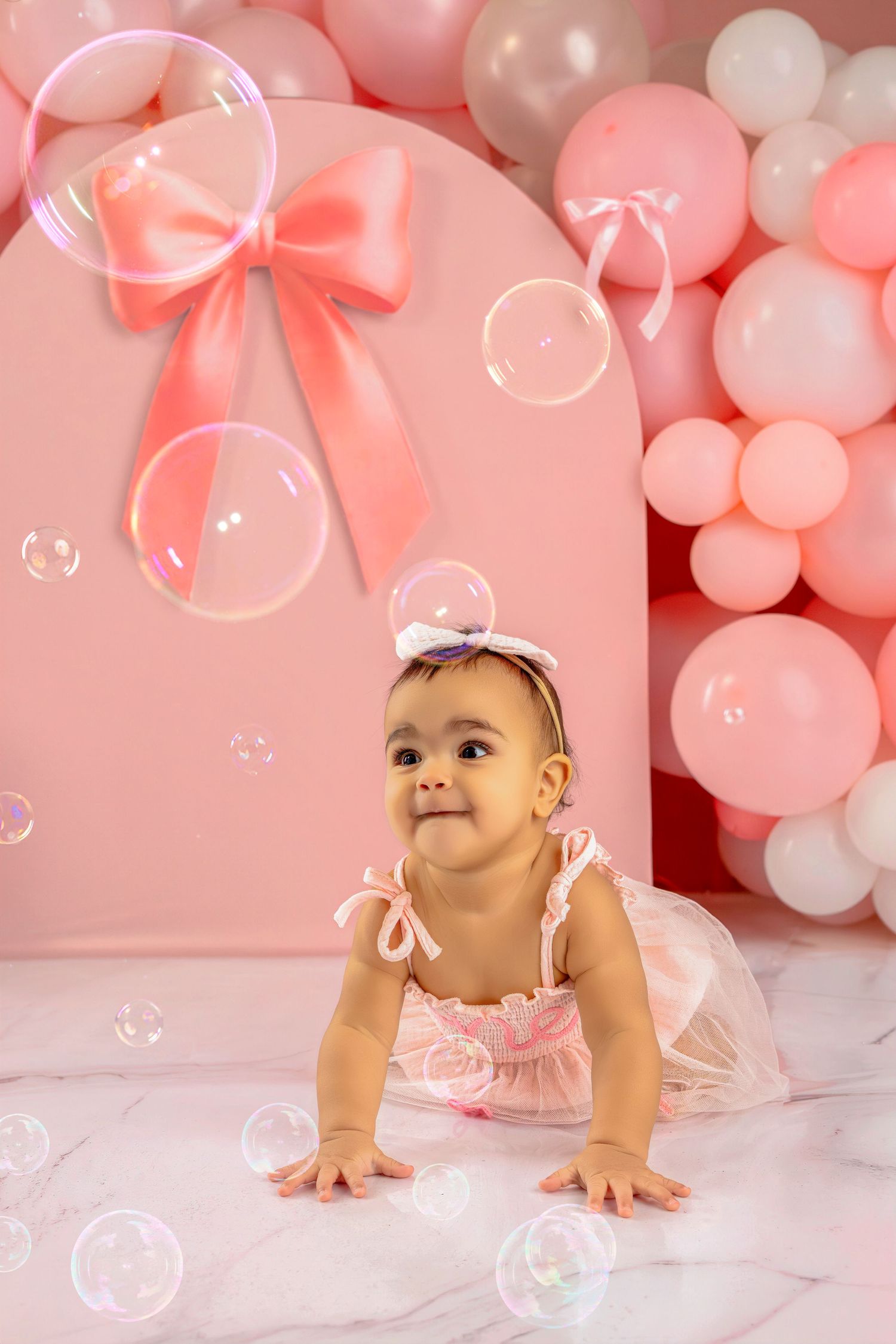 Baby crawling on white floor with pink and white balloons and bubbles in background during photo session.