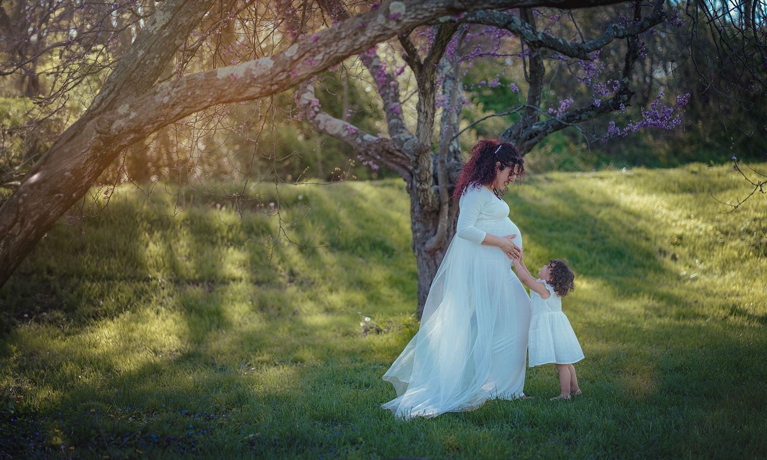Pregnant woman in white dress with toddler girl under blooming tree in sunlit garden.