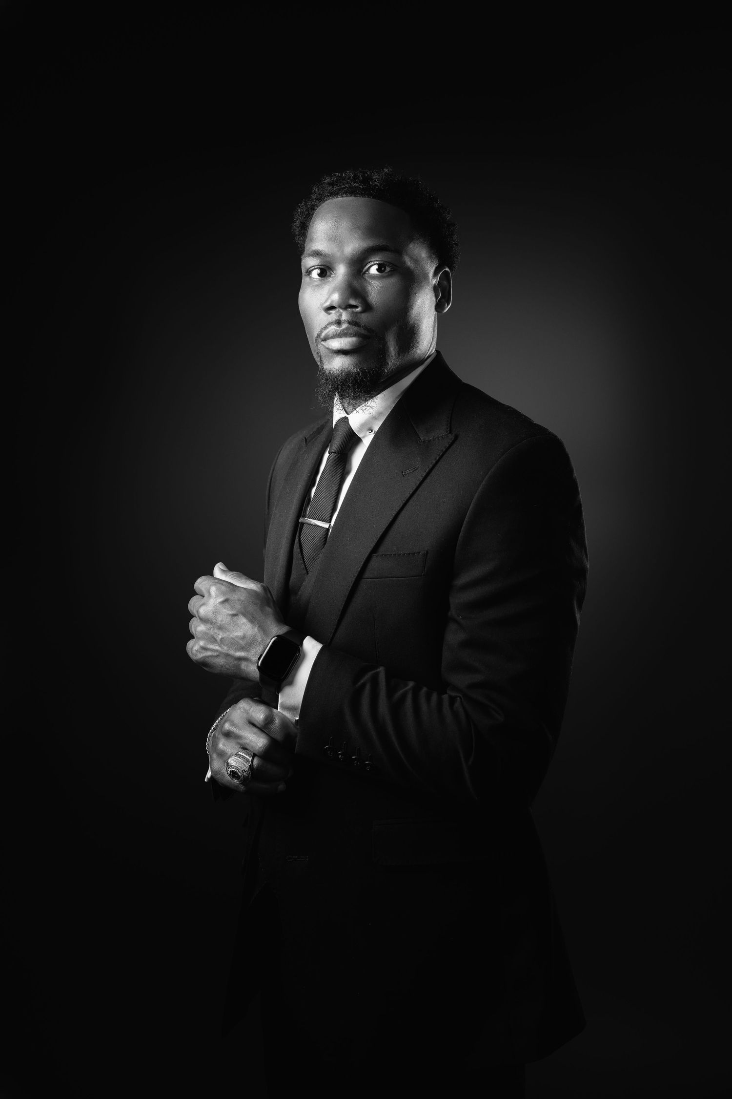 Professional black and white portrait of someone in a dark suit and tie against a dramatic shadowed background.