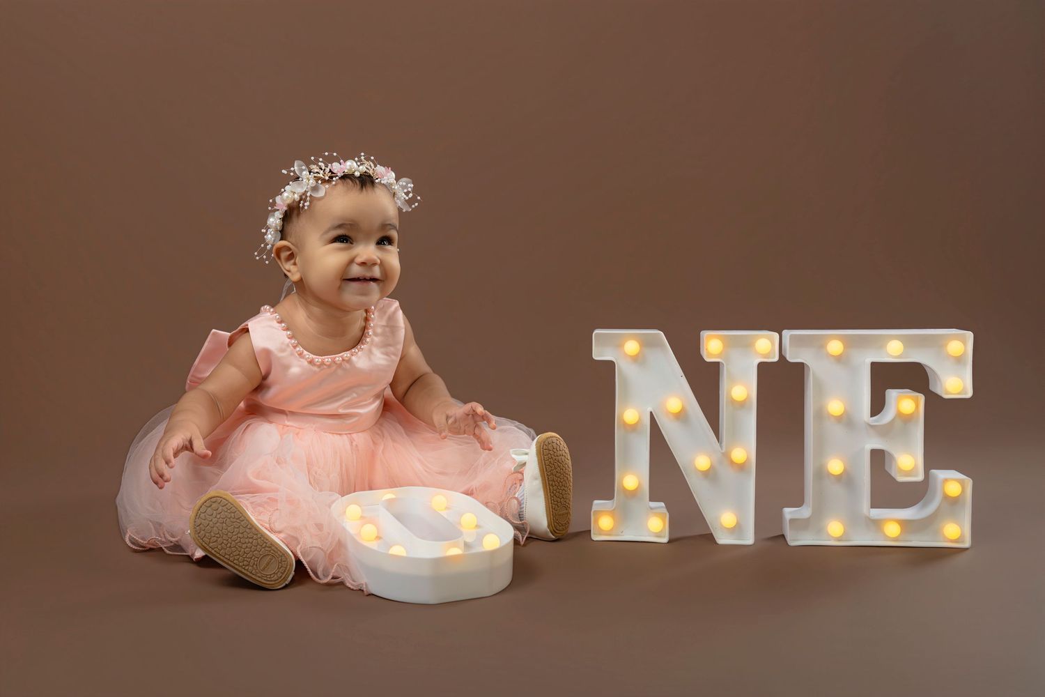 A baby in a pink dress sits next to decorative letters spelling ONE against a brown backdrop.
