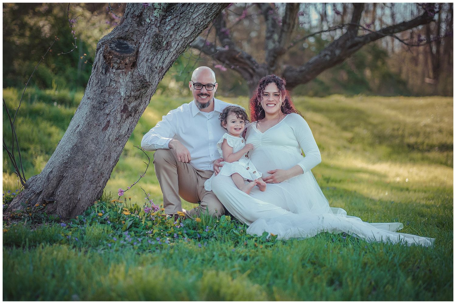 Pregnant woman in white gown poses with husband and toddler daughter under a tree on green grass for maternity photo.