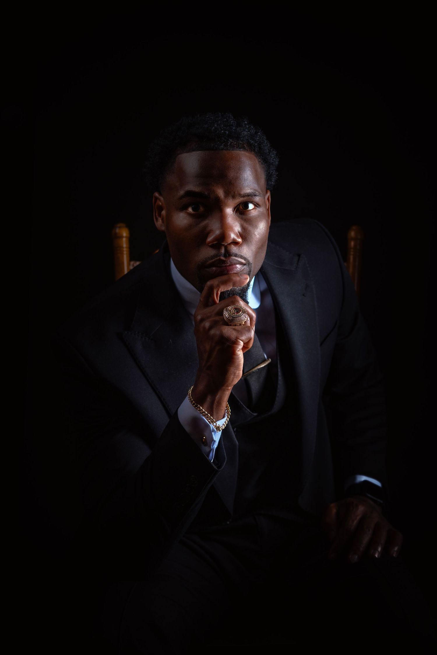 A well-dressed Black man in a dark suit sits pensively in a chair, hand raised to chin, against a black background.