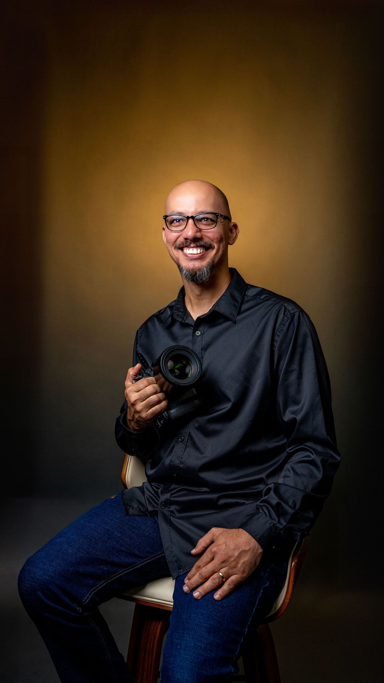 Smiling bald man with glasses holding a camera, seated on a chair in a professional studio portrait setting.