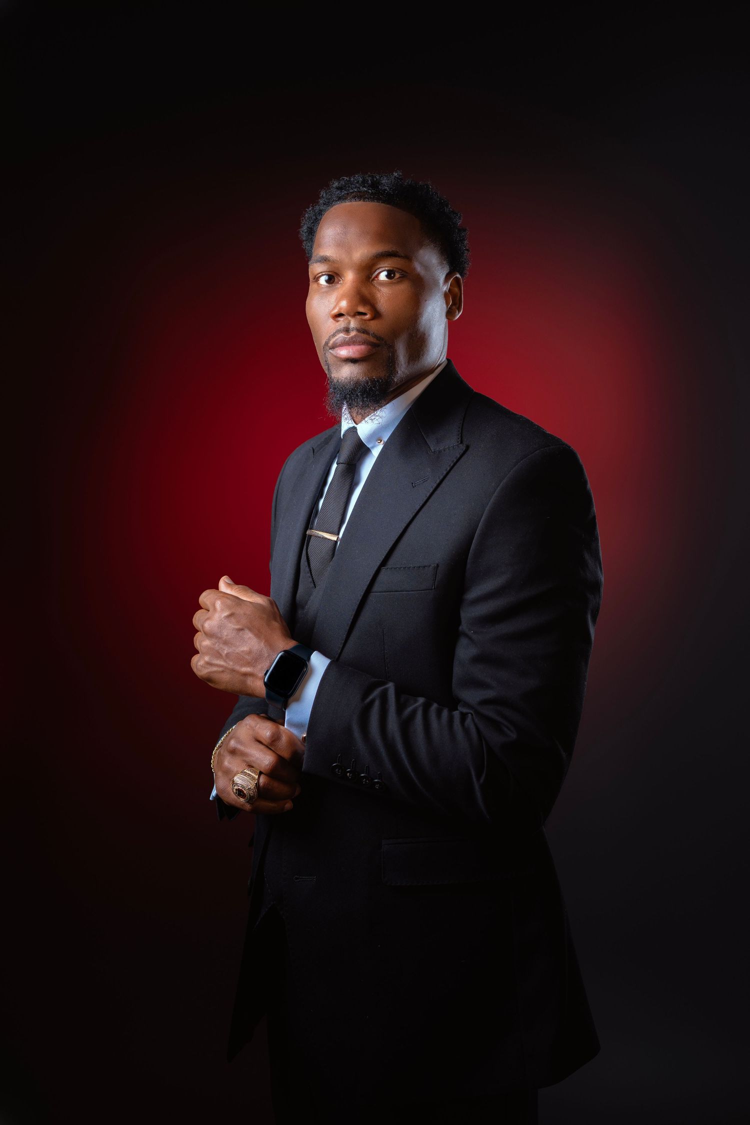 A professional Black man in a dark suit and tie poses confidently against a dramatic red and black studio background.
