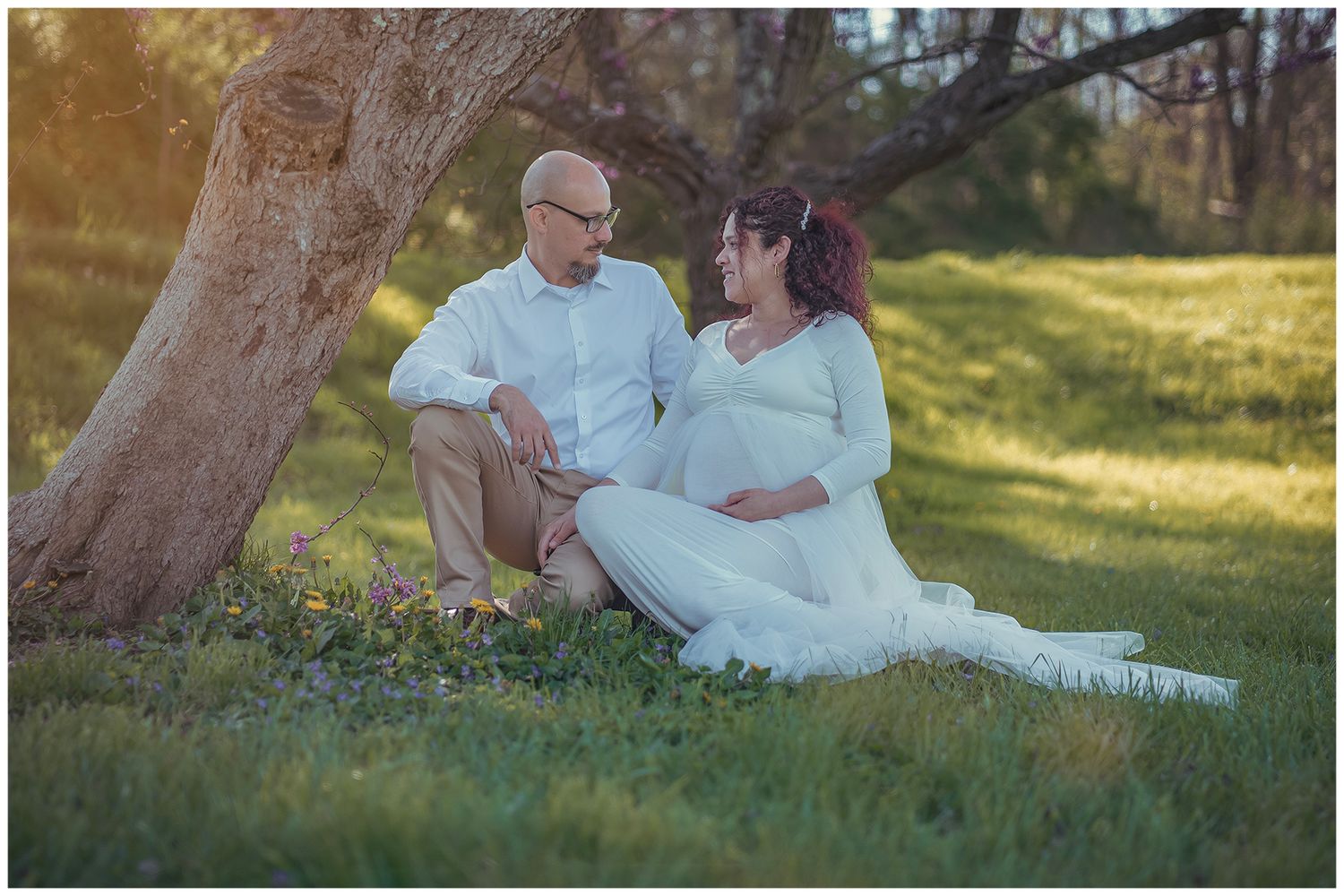 Couple sitting under a tree in a sunny park, woman in white maternity gown, man in white shirt, sharing a tender moment.