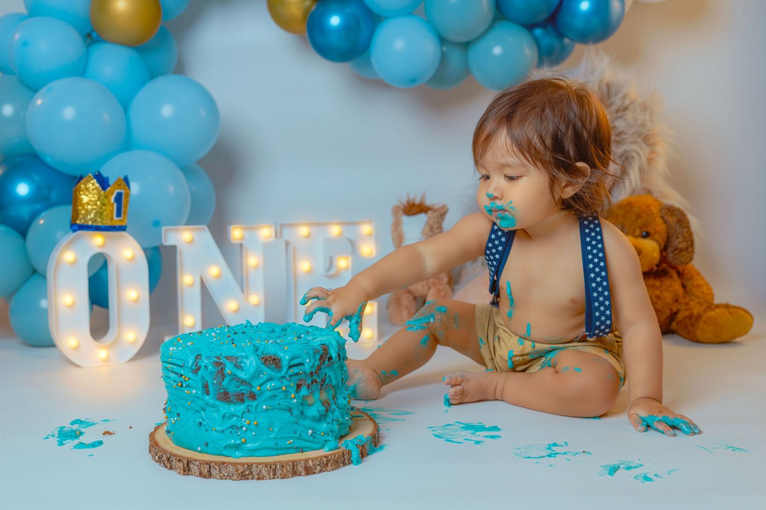 Smash cake photo session with turquoise and gold decorations featuring 'ONE' light-up letters and balloons.