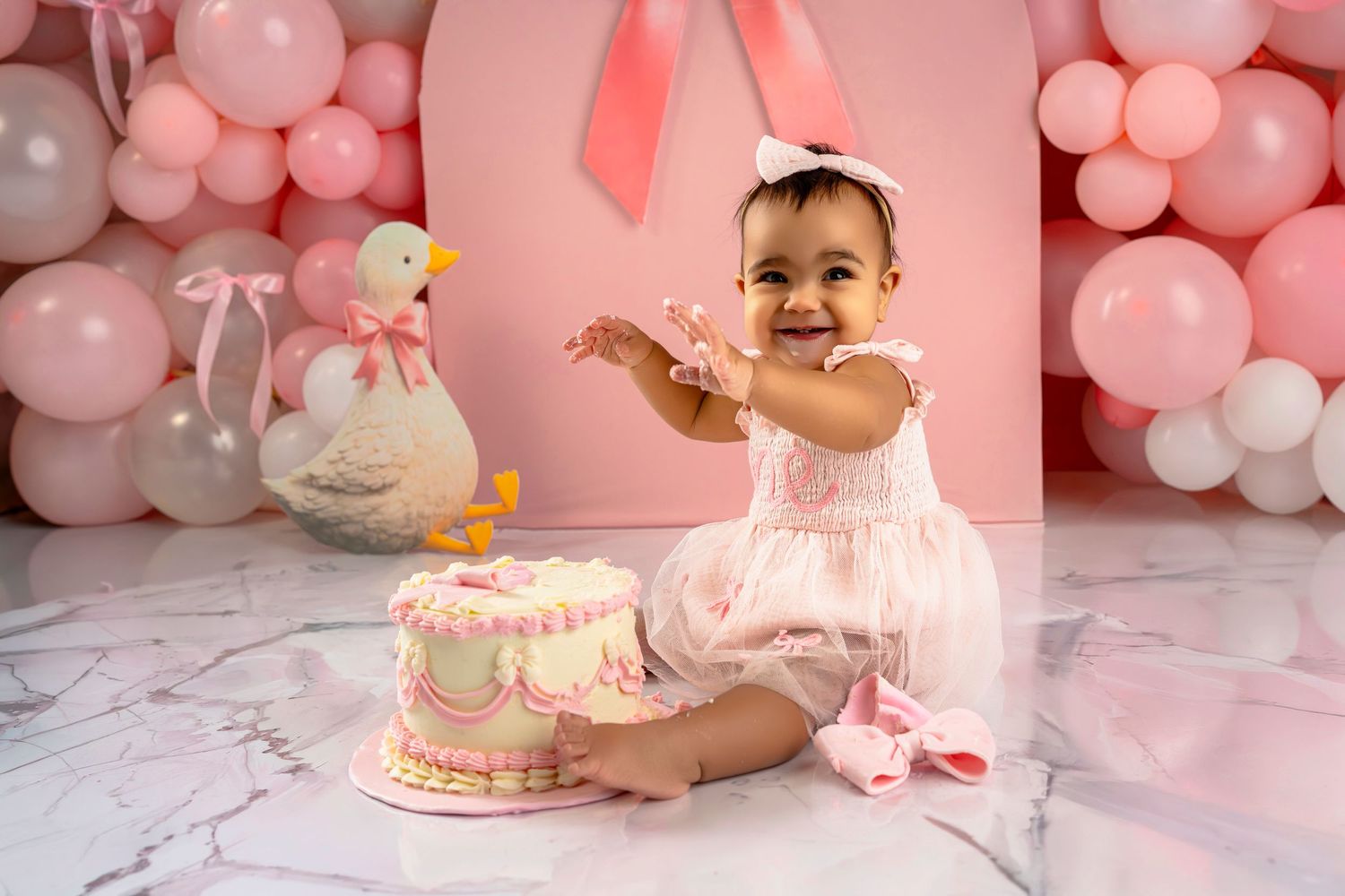 Pink and white balloon decorations create a festive backdrop for a cake smash photo session with a birthday cake and toys.