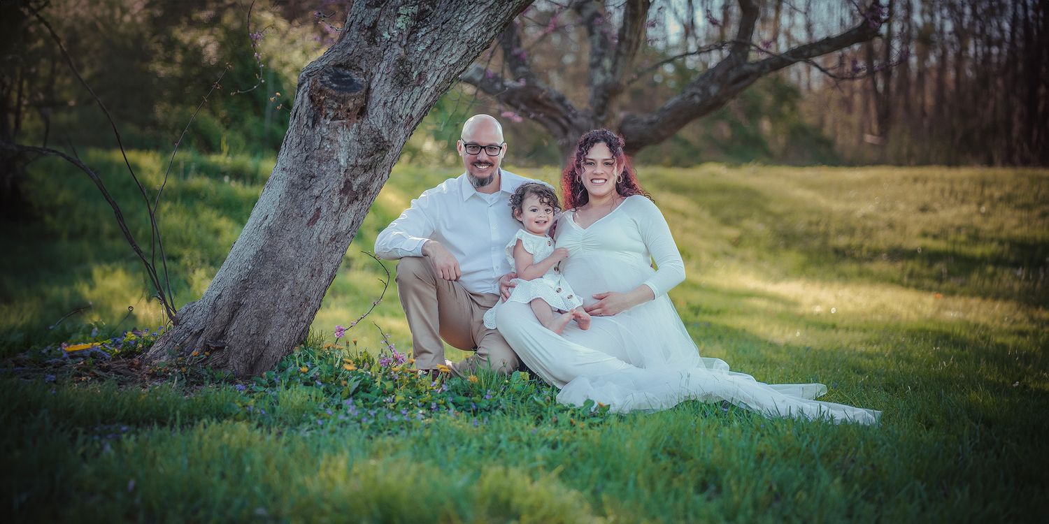 A family maternity photo session outdoors, with a pregnant woman in a white gown sitting with her husband and toddler by a tree.