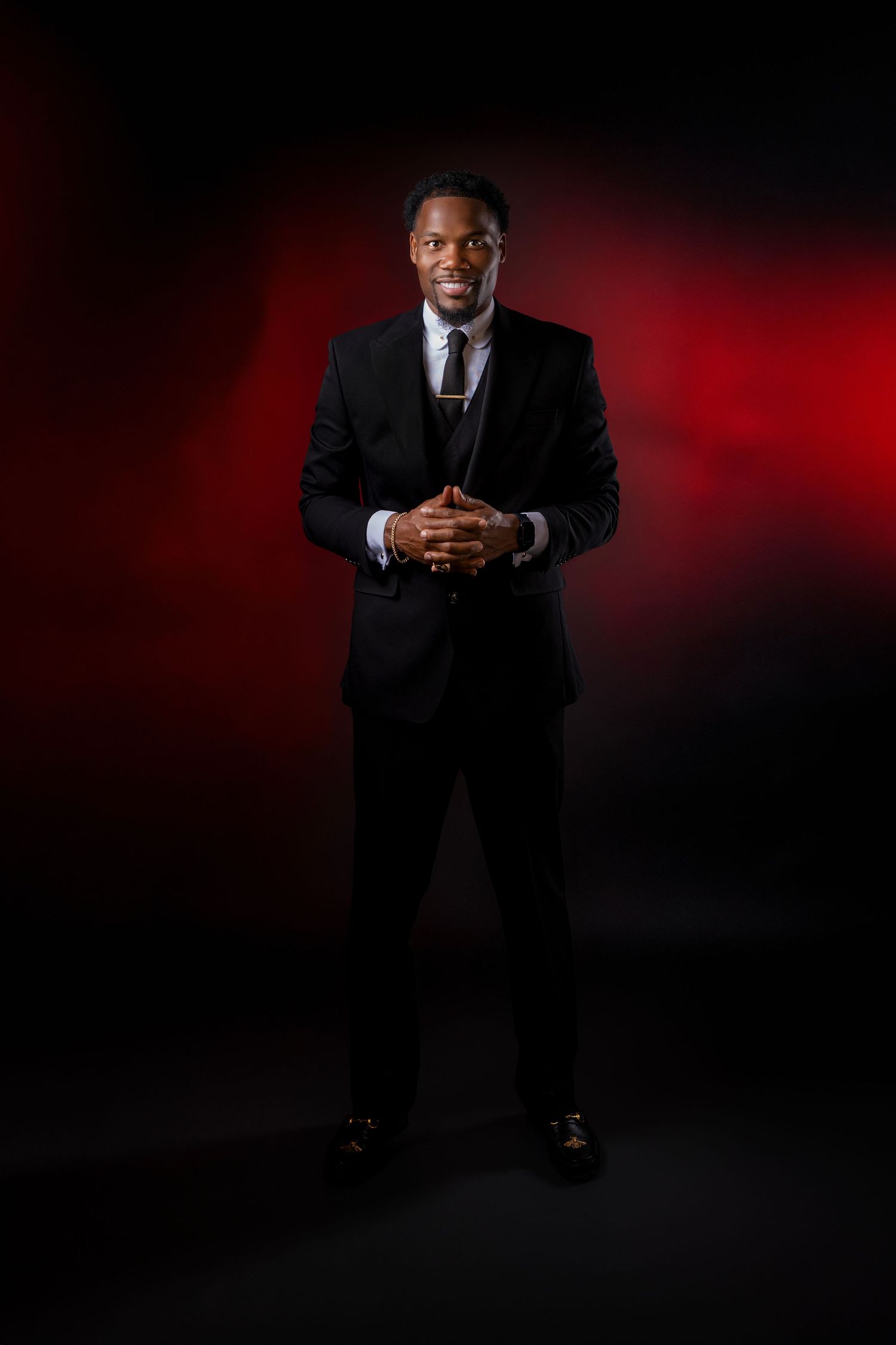 Smiling Black man in a black suit with clasped hands poses against a dark red and black studio background.