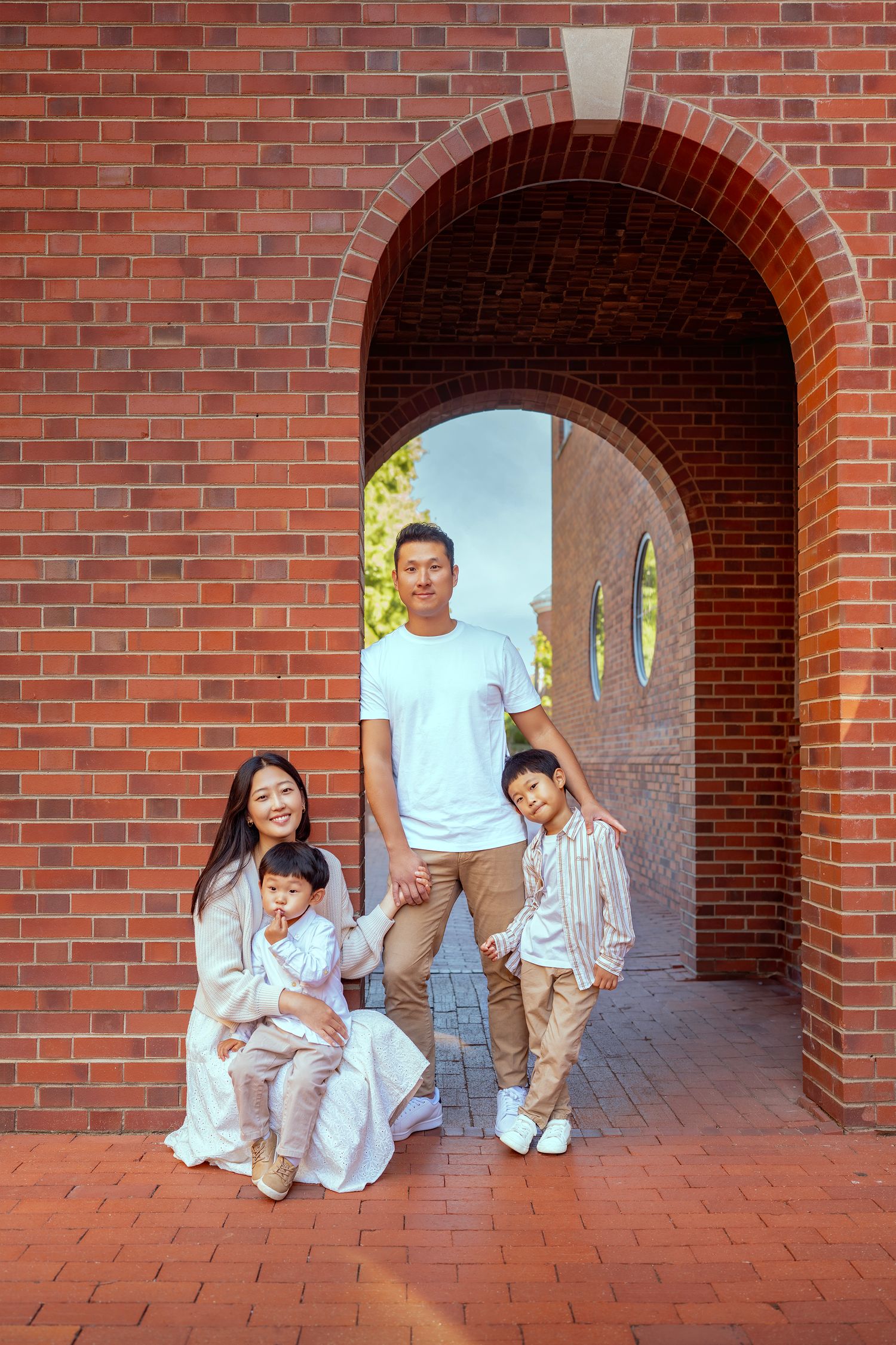 Asian family of four posing under a red brick archway, parents with two young children smiling outdoors.