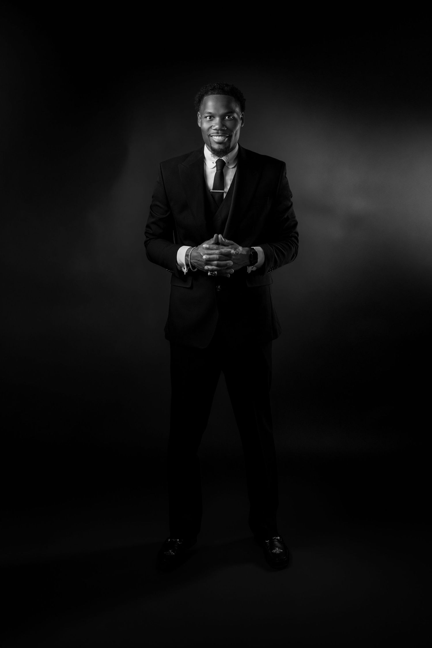A dramatic black and white portrait of someone seated in formal attire against a dark background.