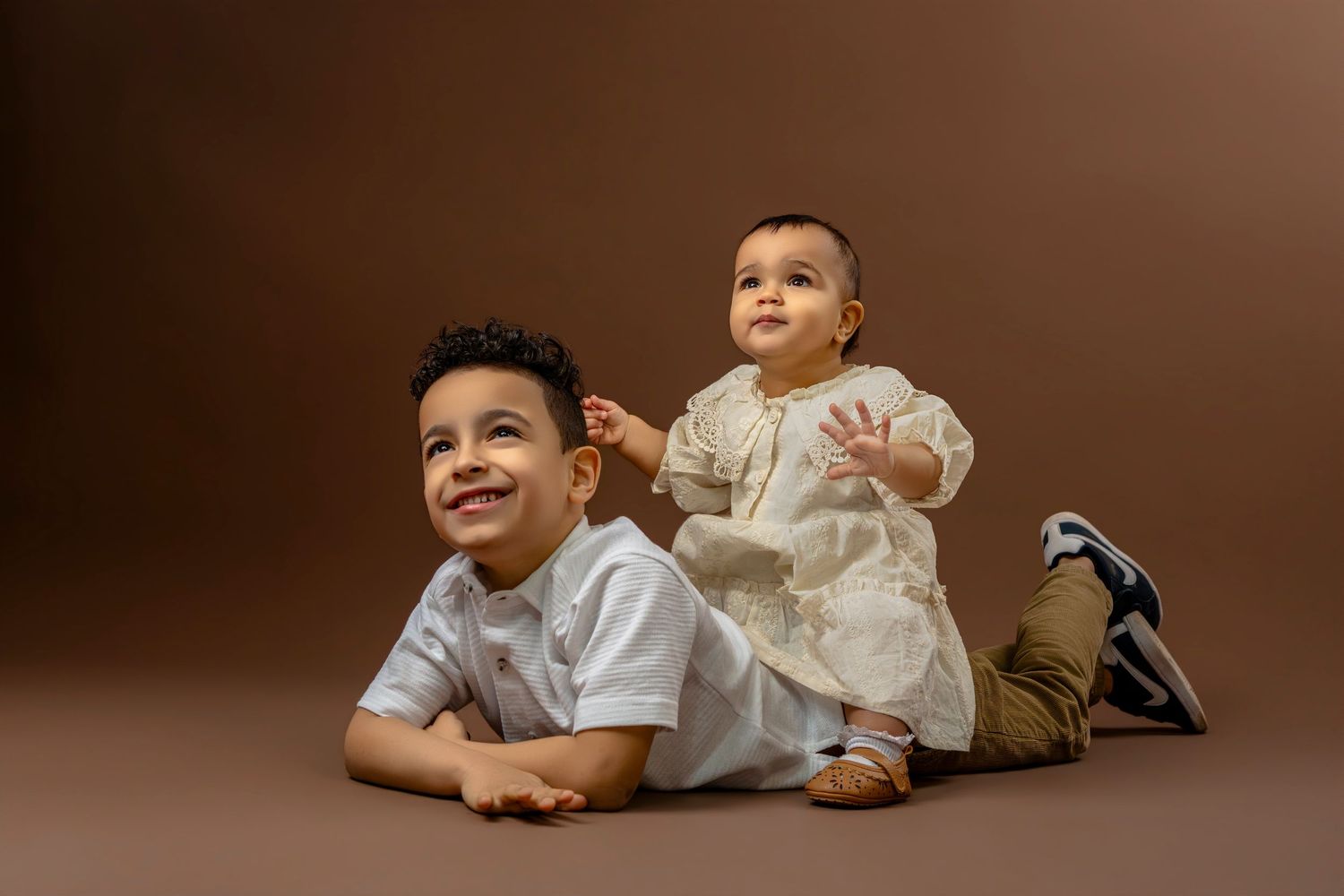 Young child in white shirt lying on brown backdrop with playful expression.