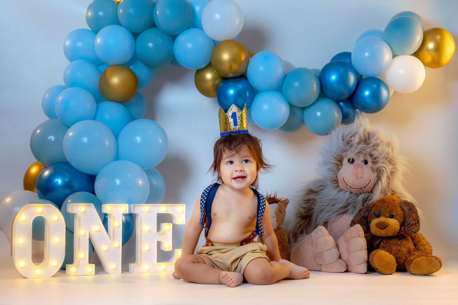 Blue and gold balloon decorations create a festive first birthday setup with stuffed animals and illuminated ONE sign.