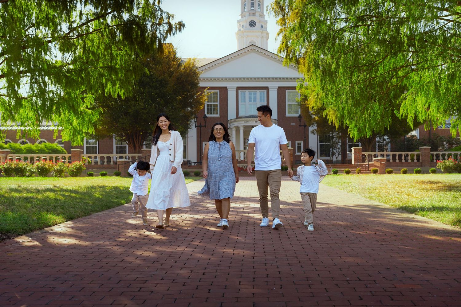 Family of five walking hand-in-hand on a brick path in front of a historic building on a sunny day.
