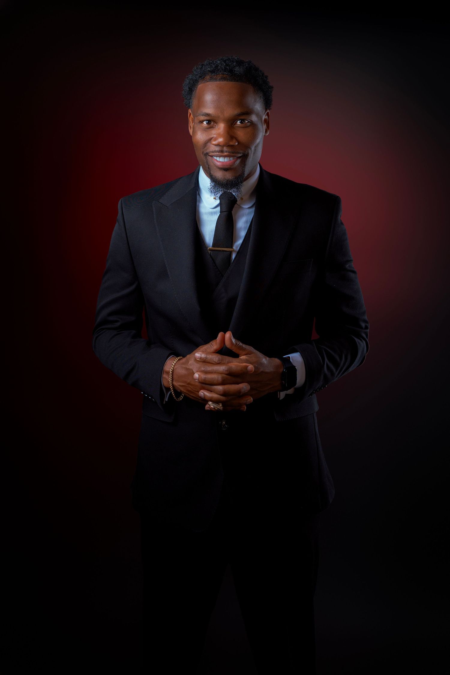 A well-dressed Black man in a black suit smiles confidently with hands clasped against a dark red background.