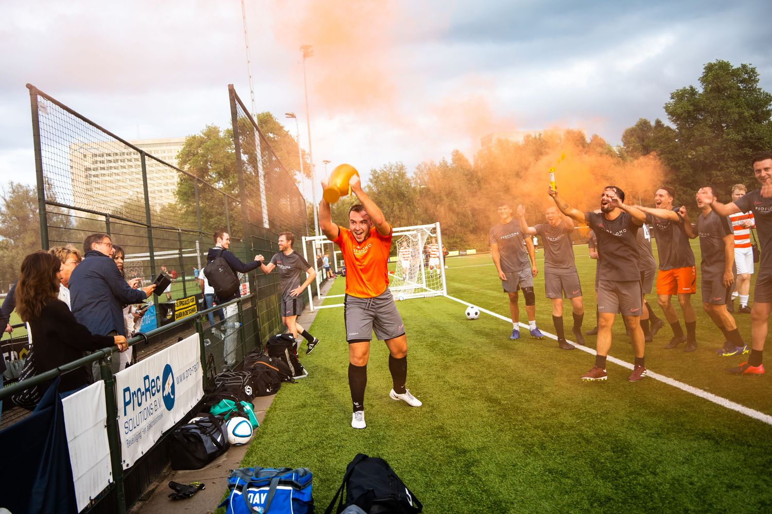 Soccer fans celebrate with orange flares and smoke during an outdoor match at sunset.