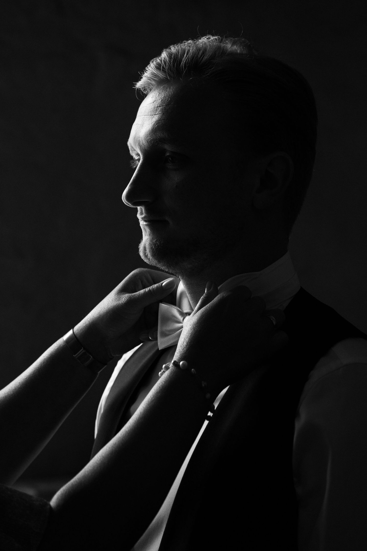 Dramatic black and white side profile portrait of a groom in formal attire against dark background.