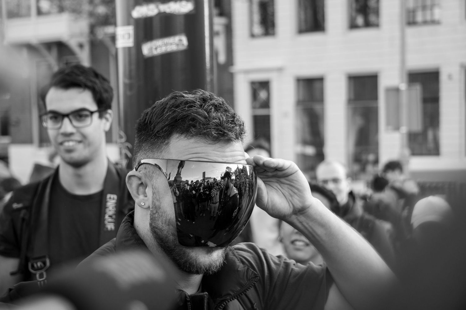 A photographer holding a reflective camera lens that mirrors the cityscape and crowd in a black and white street scene.
