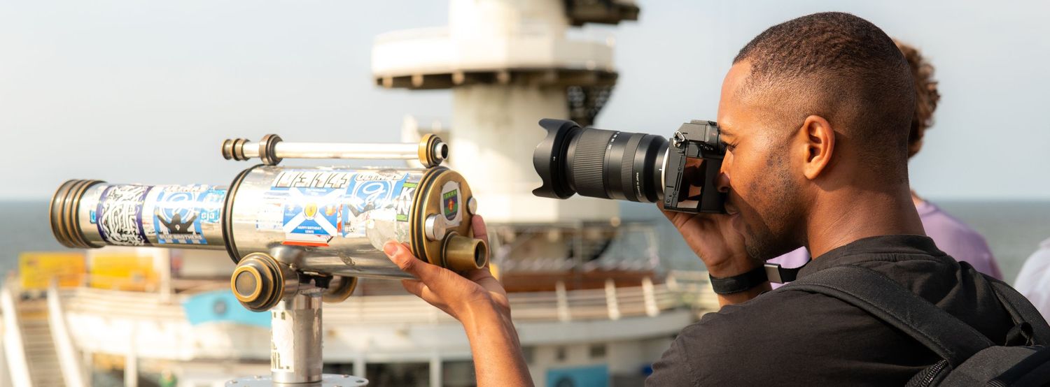 Person looking through binoculars on a cruise ship deck during golden hour with camera equipment nearby.