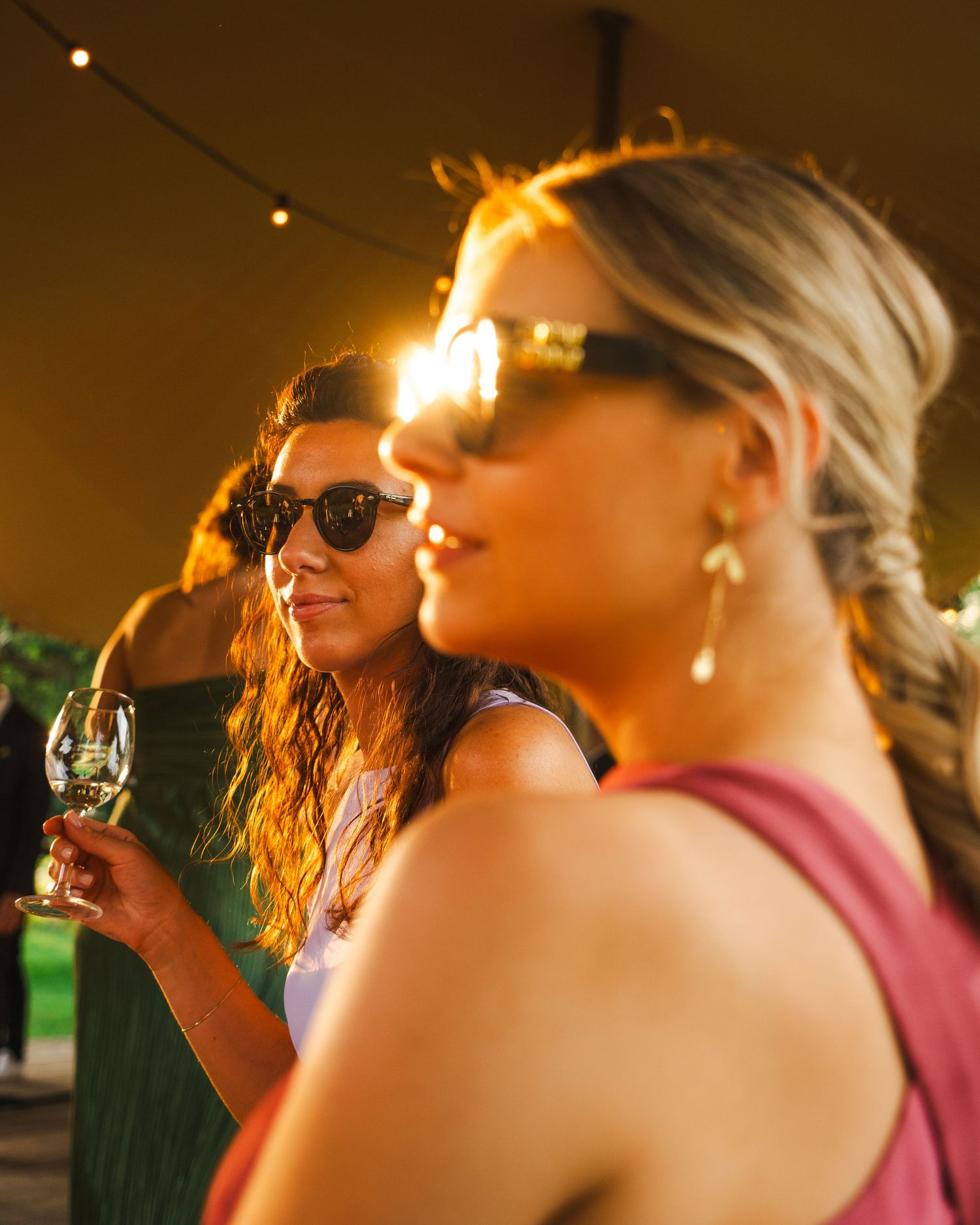 Wedding guests in sunglasses enjoying drinks during golden hour at an outdoor reception.